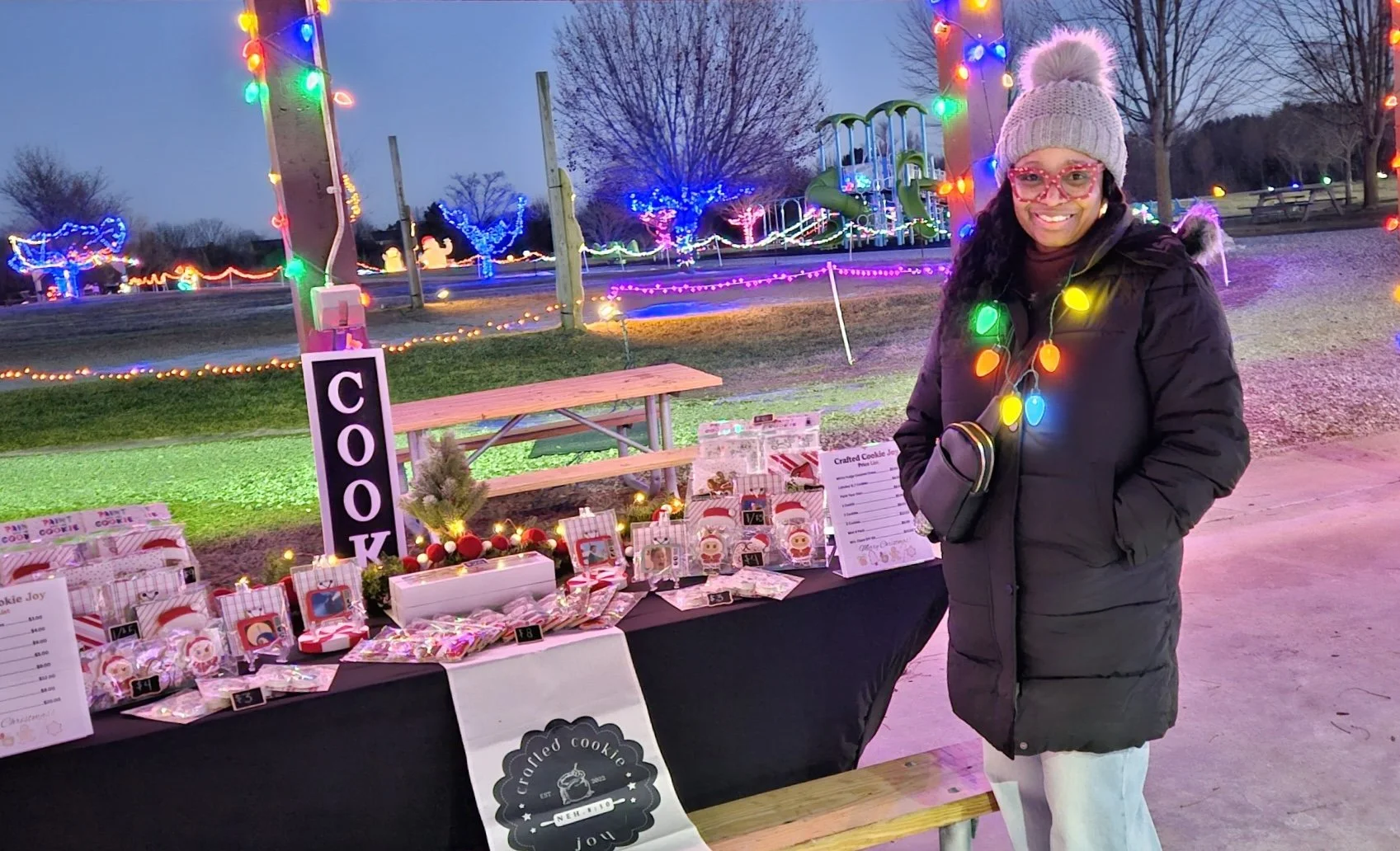 Woman in a gray knit hat and black winter coat standing next to a holiday cookie stand decorated with colorful string lights at dusk or evening.