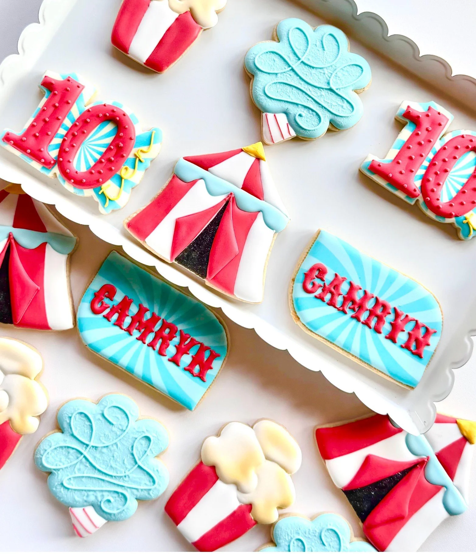 Colorful circus-themed cookies decorated with popcorn, tents, the number 10, and blue signs with red lettering reading "CAMRYN" displayed on a white tray.