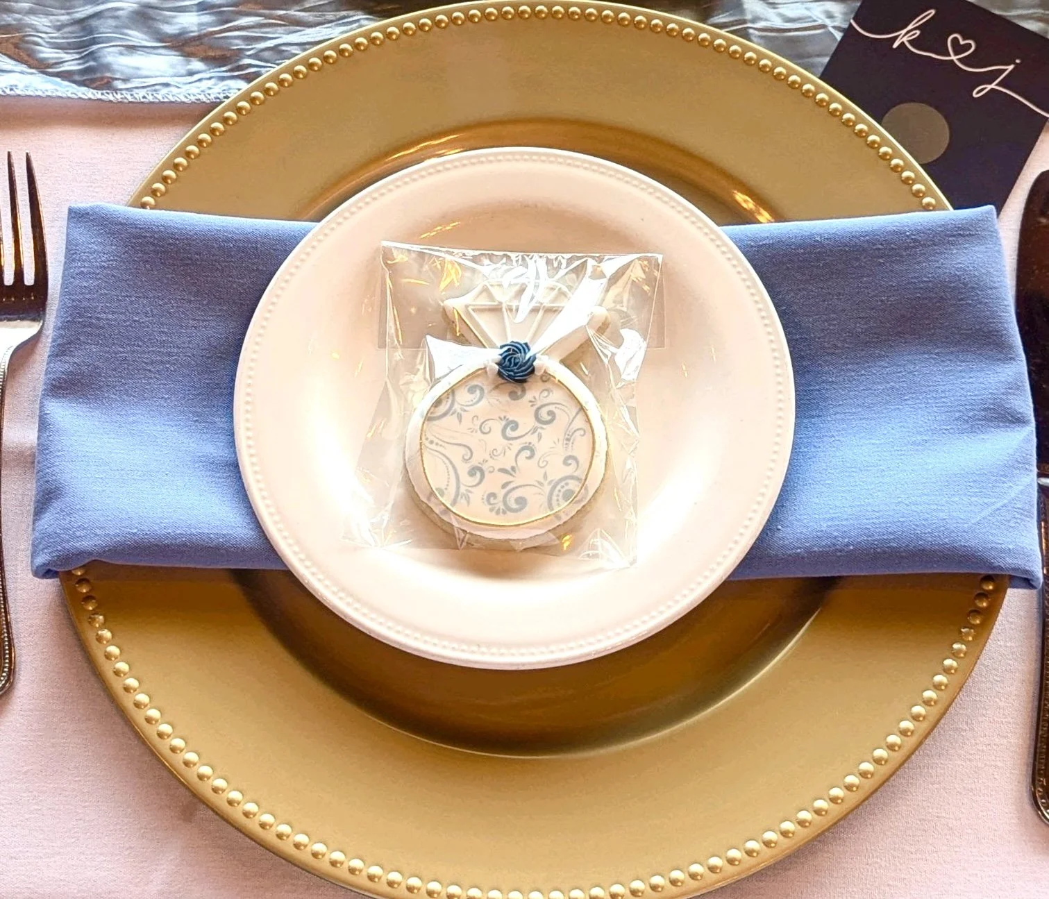A decorative table setting with a gold charger plate, a white plate with a clear plastic bag containing a decorated cookie with a blue rose and mirror, and a folded blue napkin. A black card with gold lettering and a white card with handwritten scrip