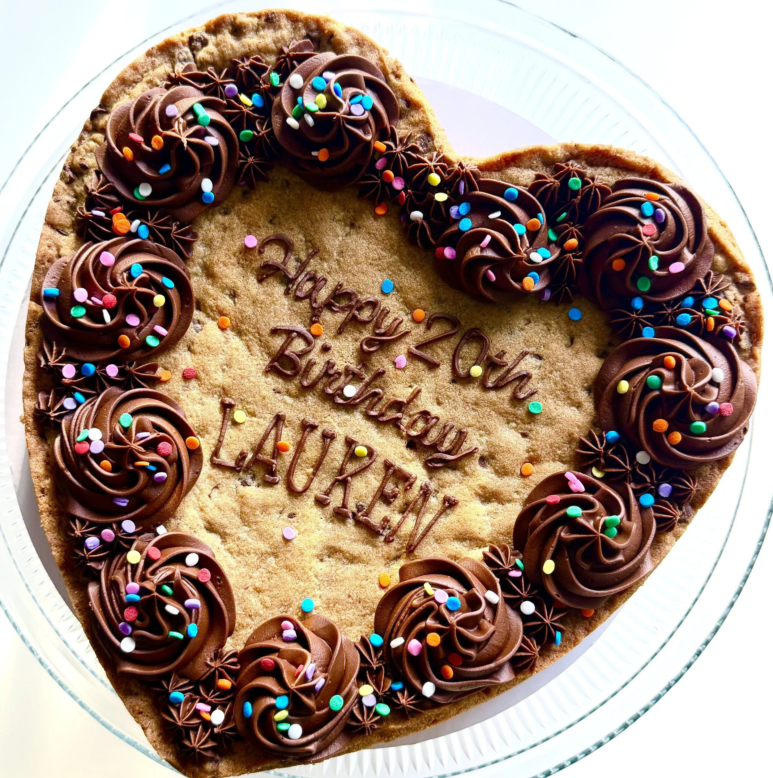 Heart-shaped cookie cake decorated with chocolate frosting and colorful sprinkles, with a handwritten message in the center that reads 'Happy 20th Birthday LAUREN'.