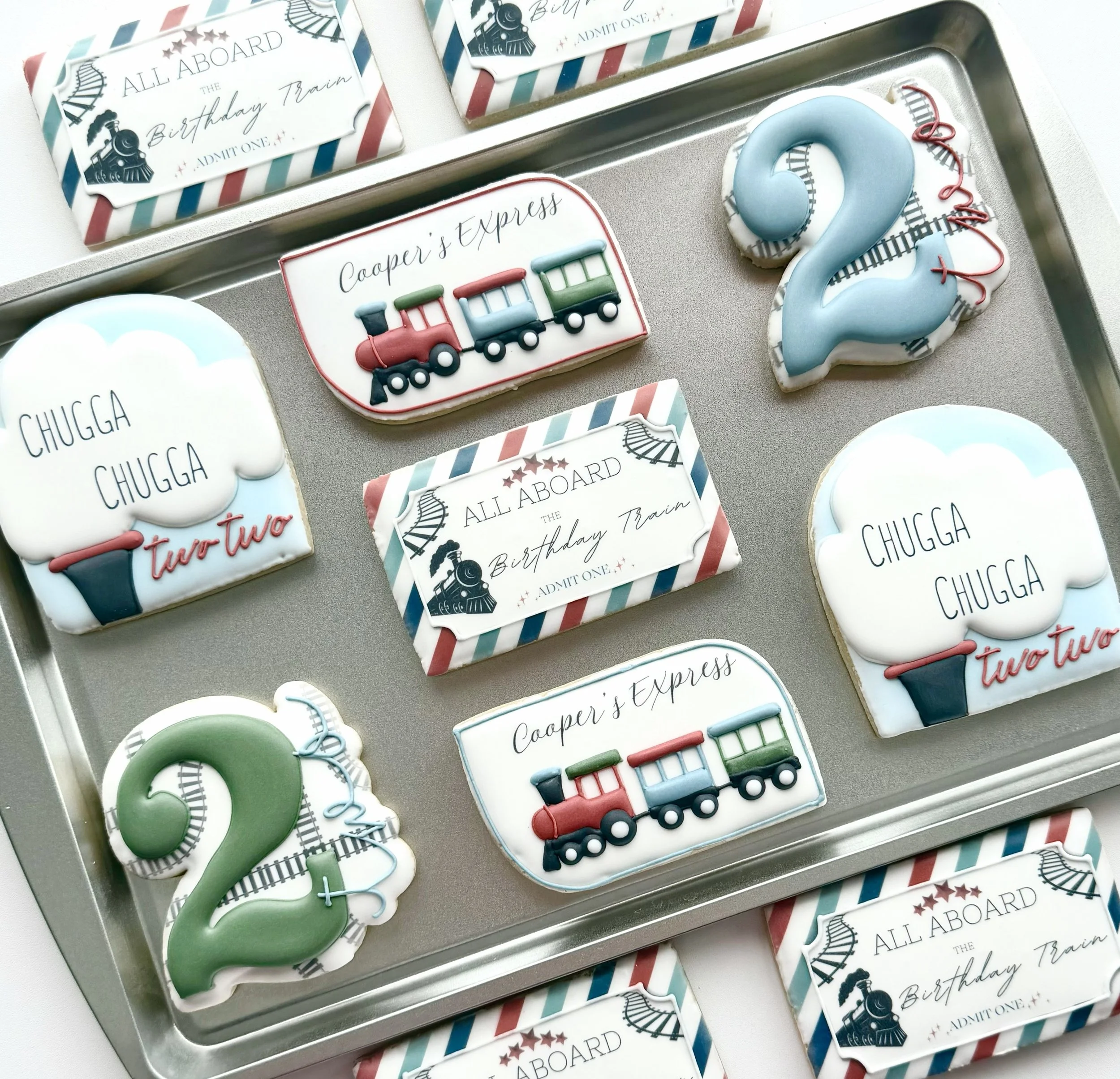 Set of decorated cookies with train theme for a birthday party, featuring numbered candles for age 2, train illustrations, and birthday messages like "All Aboard the Birthday Train" and "Chugga Chugga two two."