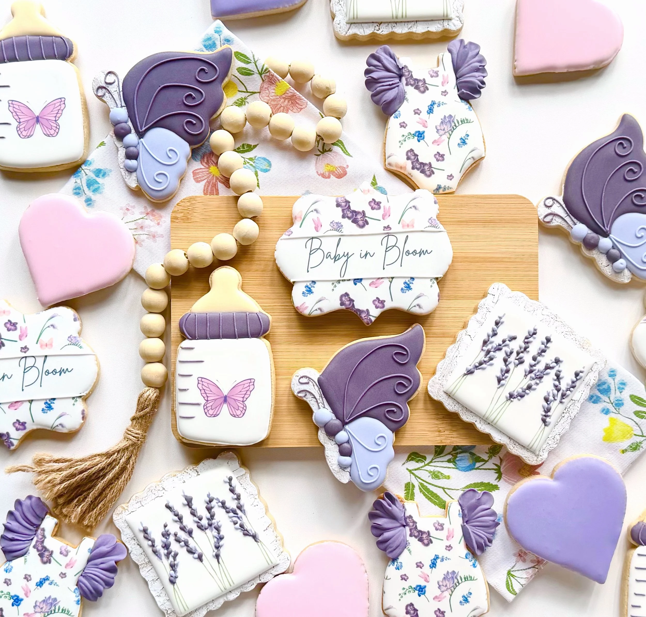 Assorted decorated cookies and a beaded banner with a baby shower theme, featuring butterfly and baby bottle designs, floral patterns, and a central sign that reads "Baby in Bloom."
