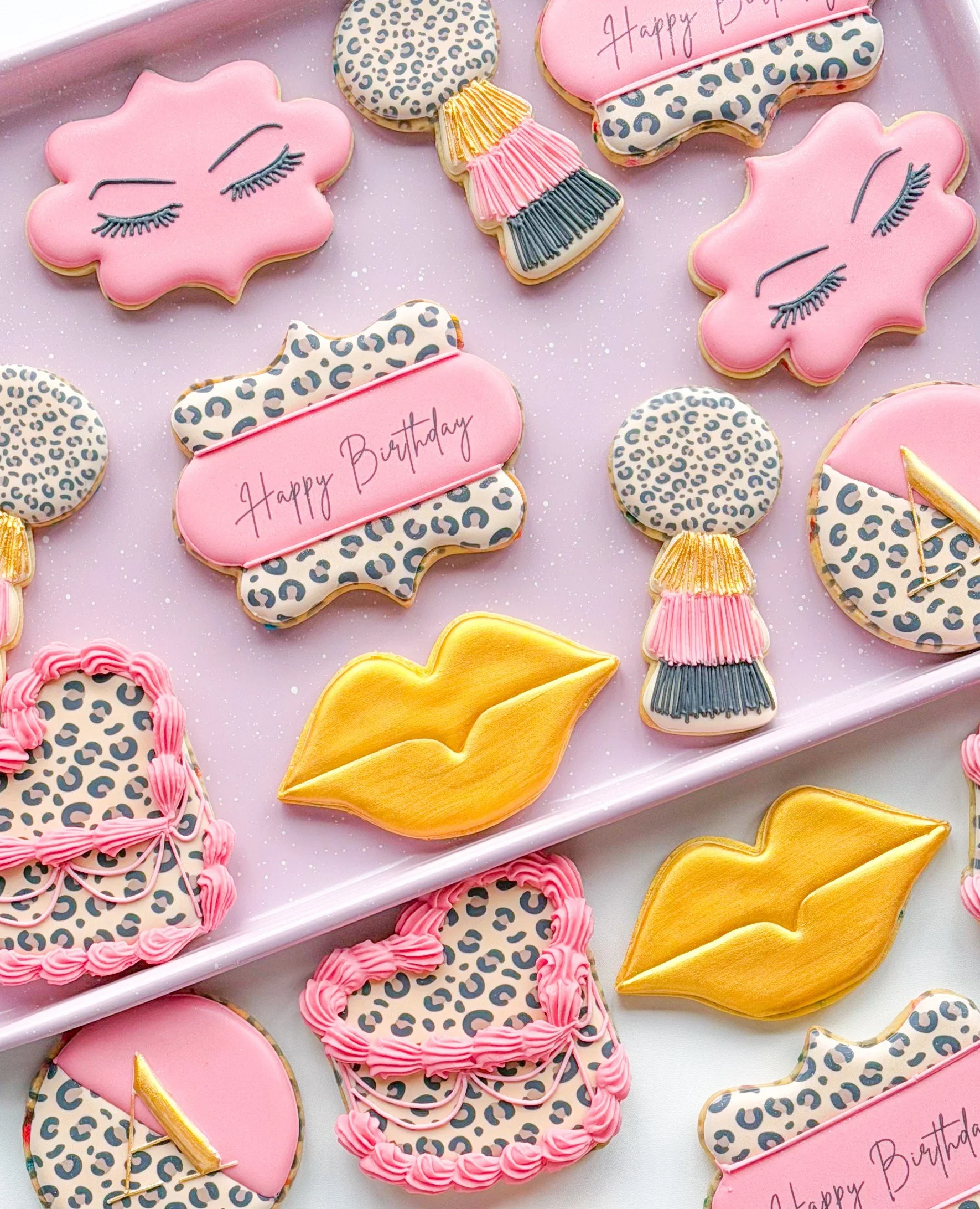 Assorted decorated birthday cookies with pink, gold, and leopard print designs on a pink and white background.