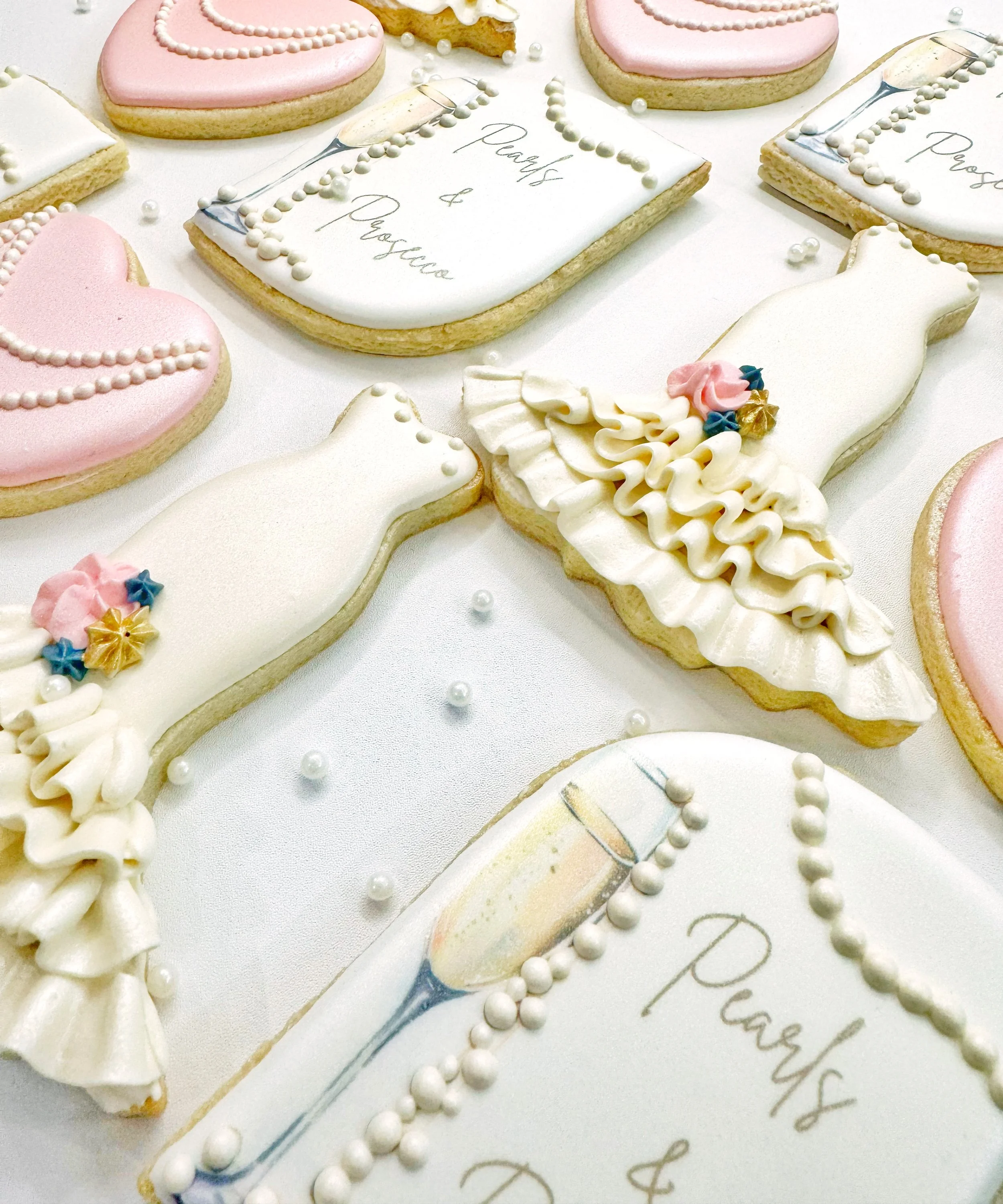Decorated cookies themed for a bachelorette or bridal celebration, including hearts, champagne glasses, and a woman in a dress, with pastel pink, white, and gold accents, and edible pearl decorations.