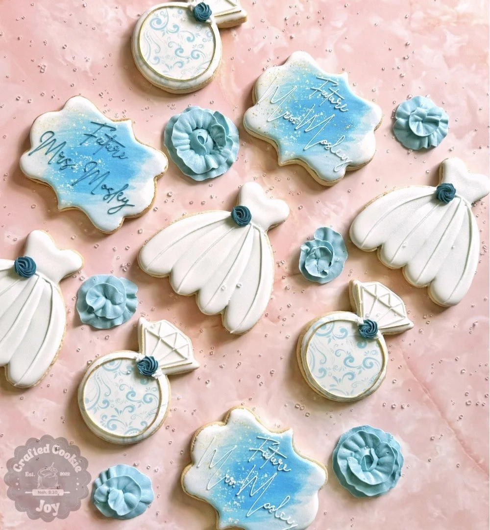 Decorated cookies on a pink surface, including a wedding dress, engagement ring, and plaque-shaped cookies with blue and white icing, and small blue flowers.