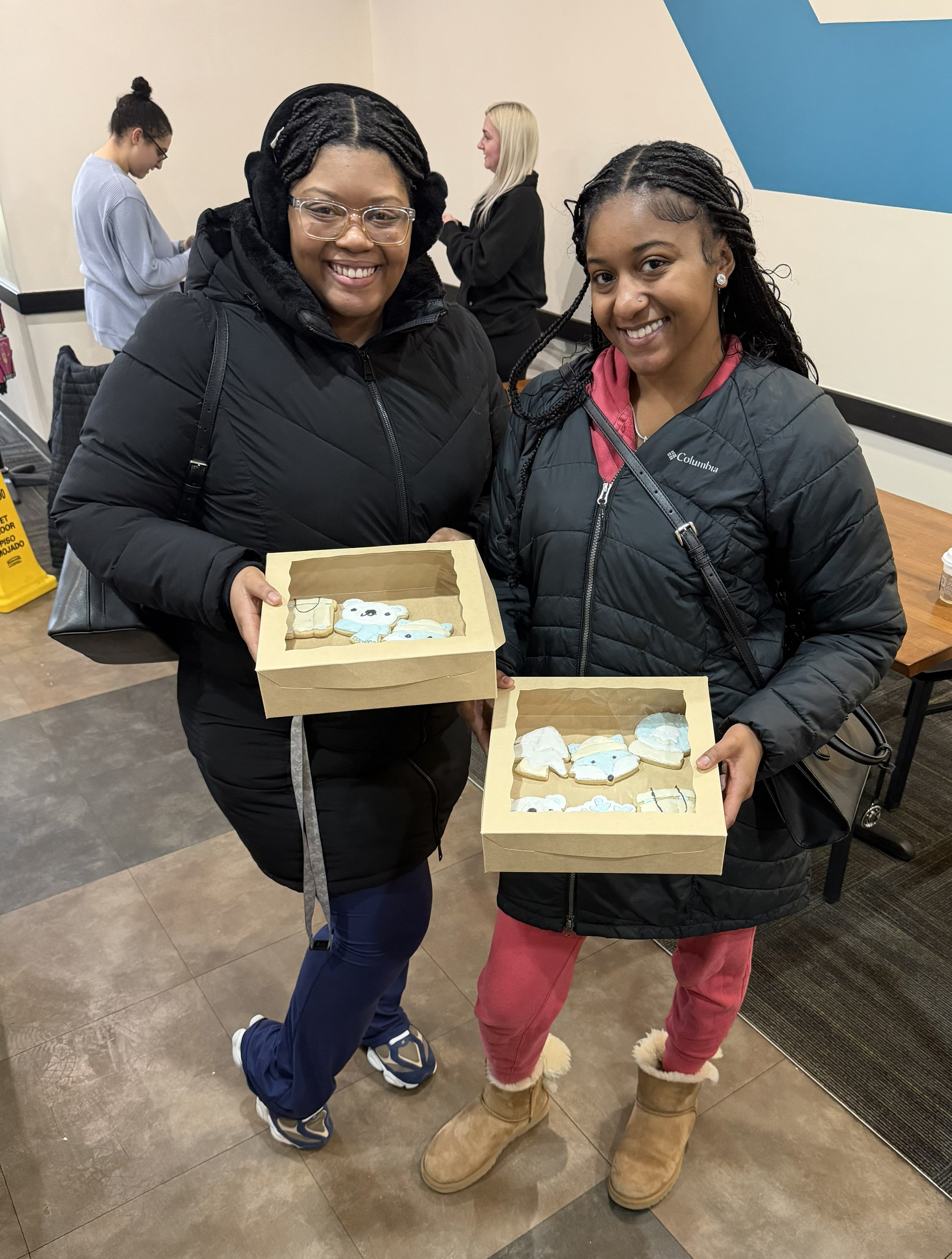 Two women wearing warm jackets smiling and holding boxes of decorated cookies shaped like snowmen, snowflakes, and winter themes inside a room.