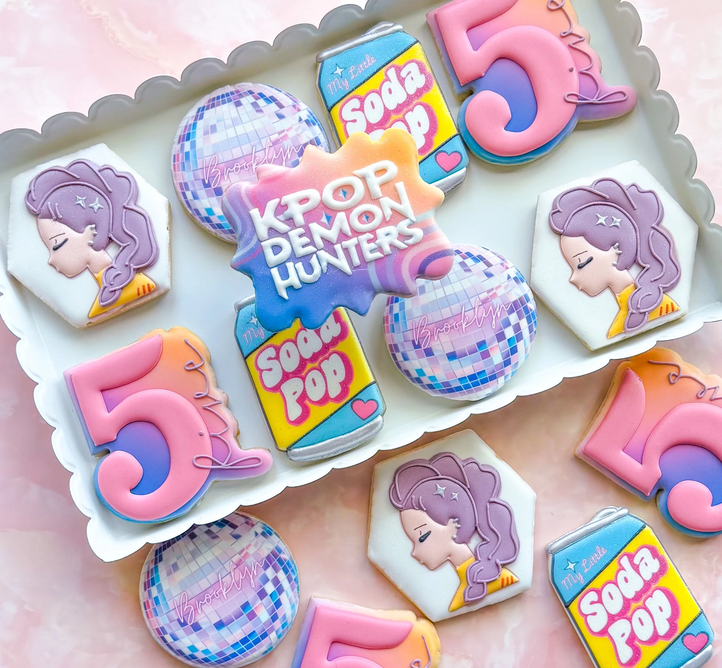 Colorful cookies decorated with pastel icing featuring characters from Kpop, a disco ball cookie, "kpop demon hunters" plaque cookie, and the number 5, and sugar cookies shaped like soda pop can with pink and purple accents.
