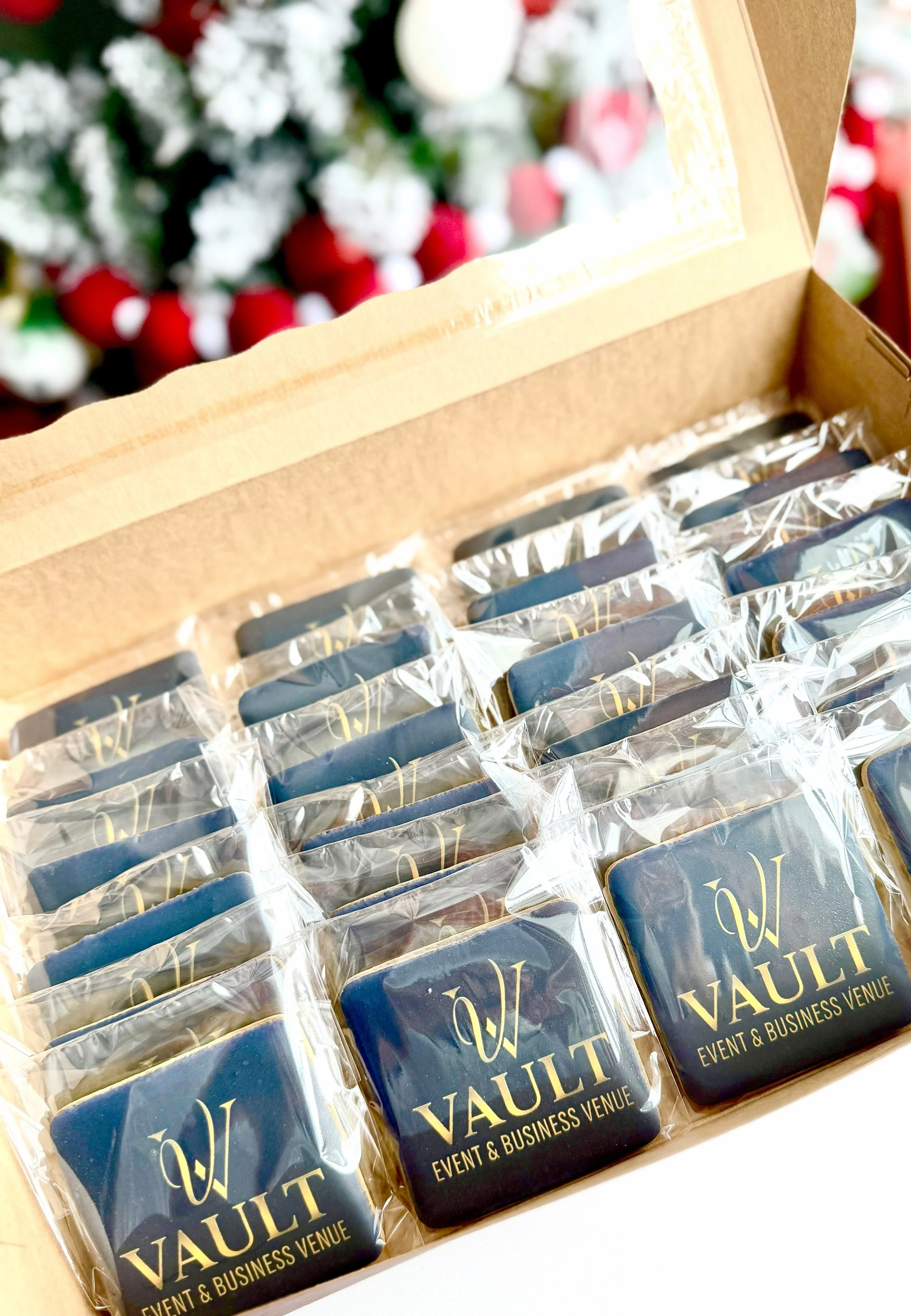 A box of individually wrapped cookies with the logo 'Vault Event & Business Venue' on each, placed on a table with a blurred background of Christmas decorations.