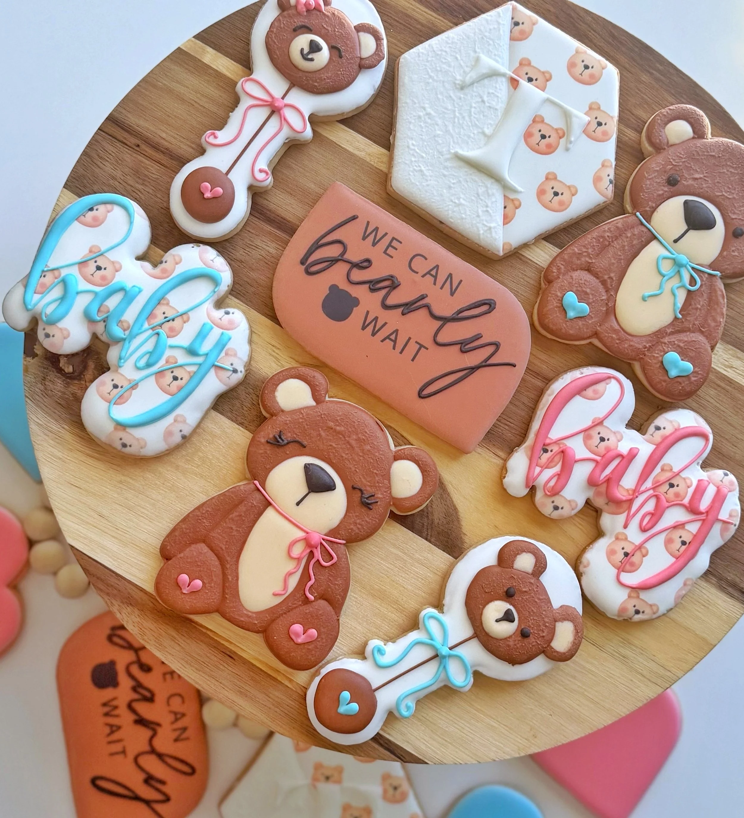 Assorted bear-shaped cookies with different decorations, a brown plaque with the message 'We can bearly wait,' and a white hexagon shaped cookie with bear faces on top, all arranged on a wooden tray.