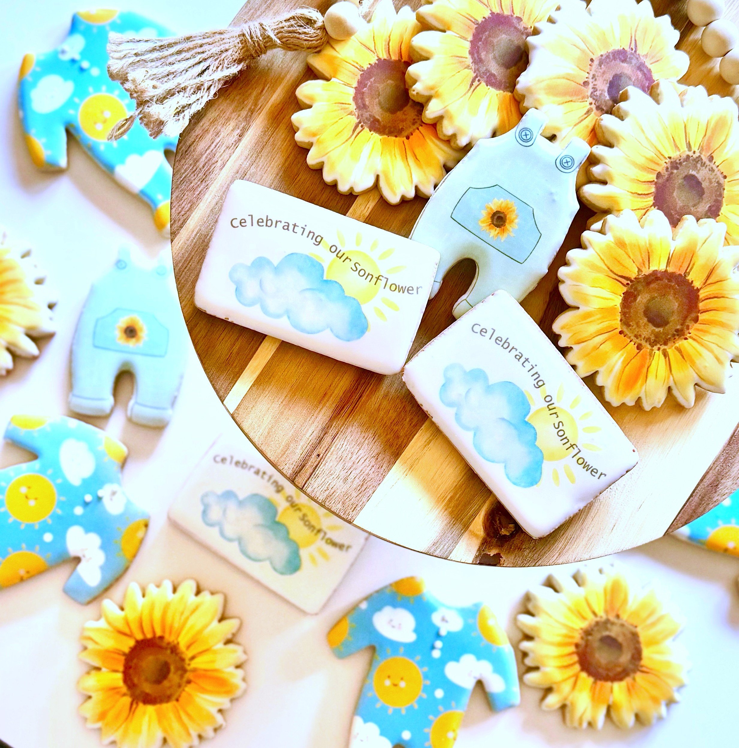 Decorative cookies on a wooden platter. The cookies include sunflower-themed designs, clouds with the text "celebrating our sonflower," and sunflower-patterned clothing cookies.