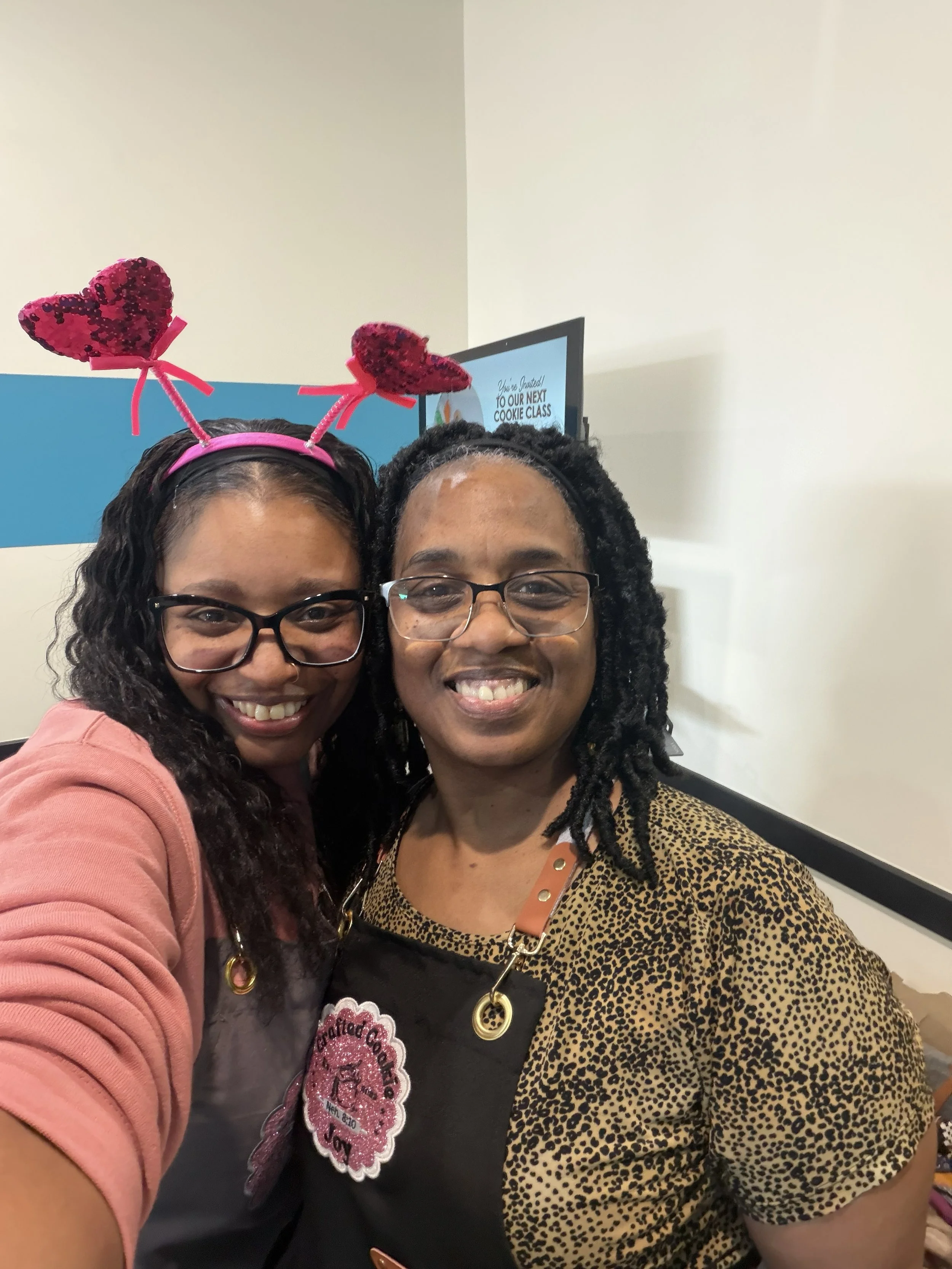 Two women smiling for a selfie, one wearing heart-shaped headbands with red sequins, and both wearing aprons with festive patches, standing in a room with a TV in the background.
