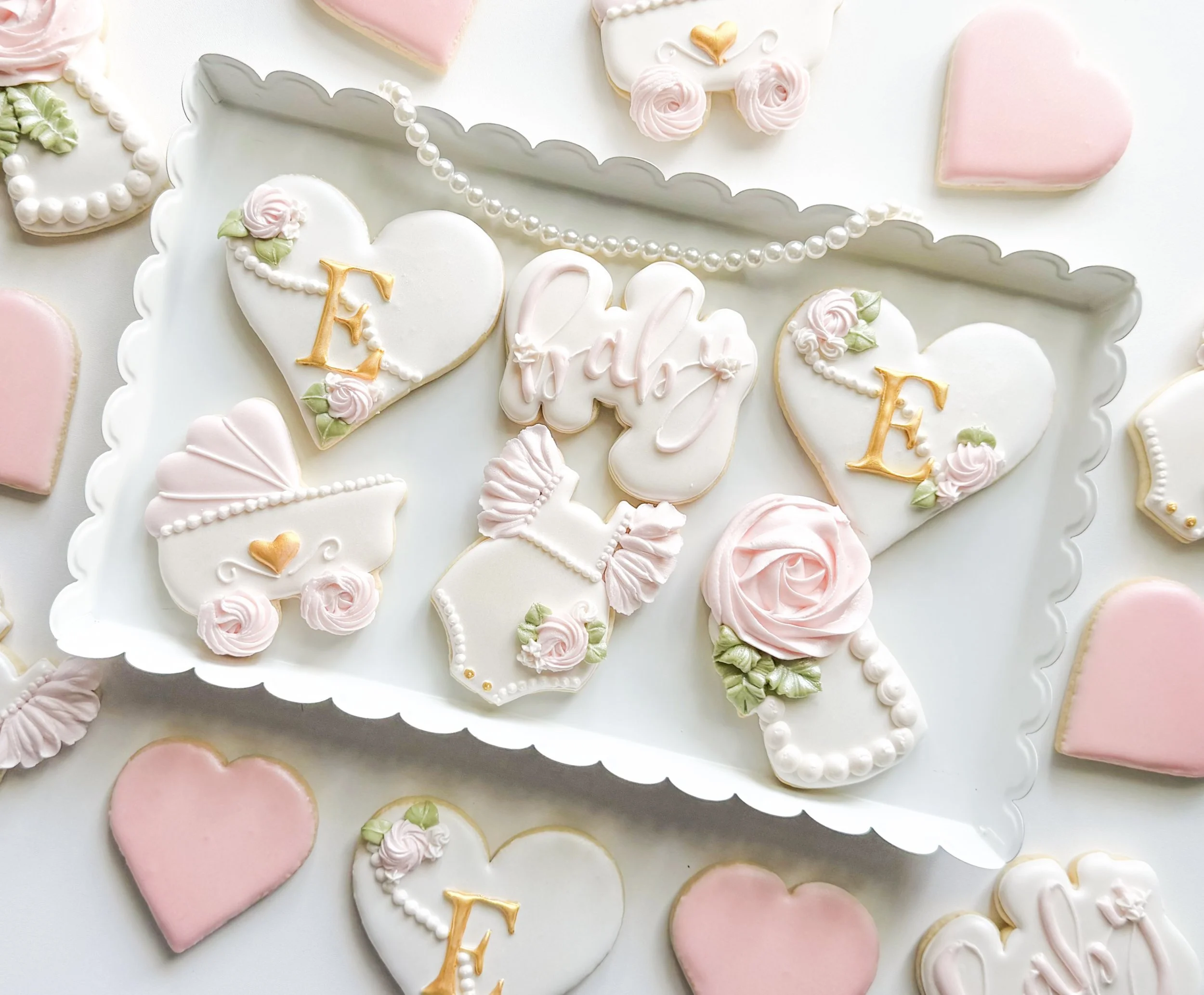 Close-up of decorated cookies on a white tray with pink, white, and pastel colors featuring hearts, roses, and the word  'baby' in gold lettering for a celebration or special occasion.