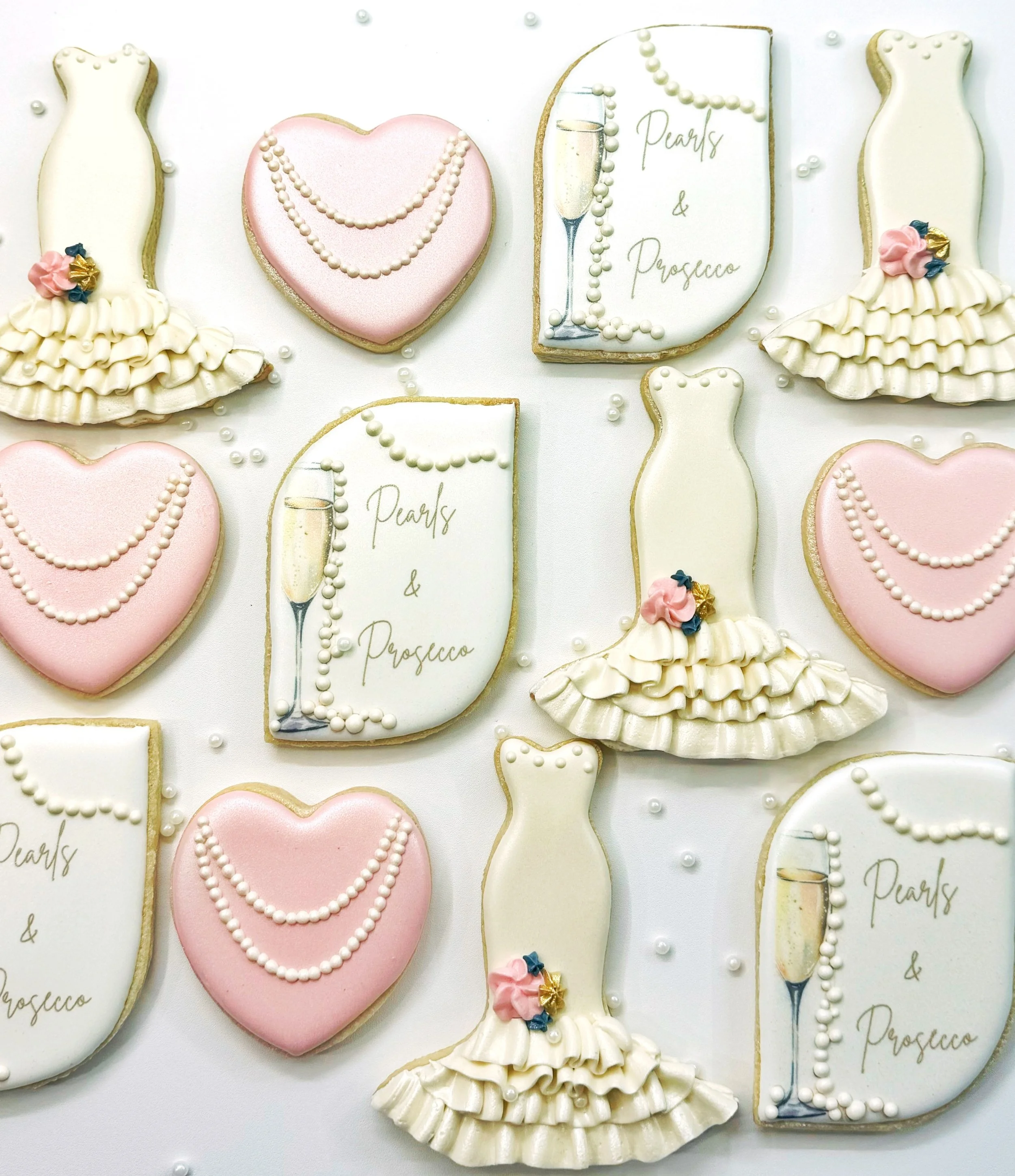 Decorative cookies shaped like wedding dresses, pink hearts, and champagne glasses, with white icing and floral details on a white background.