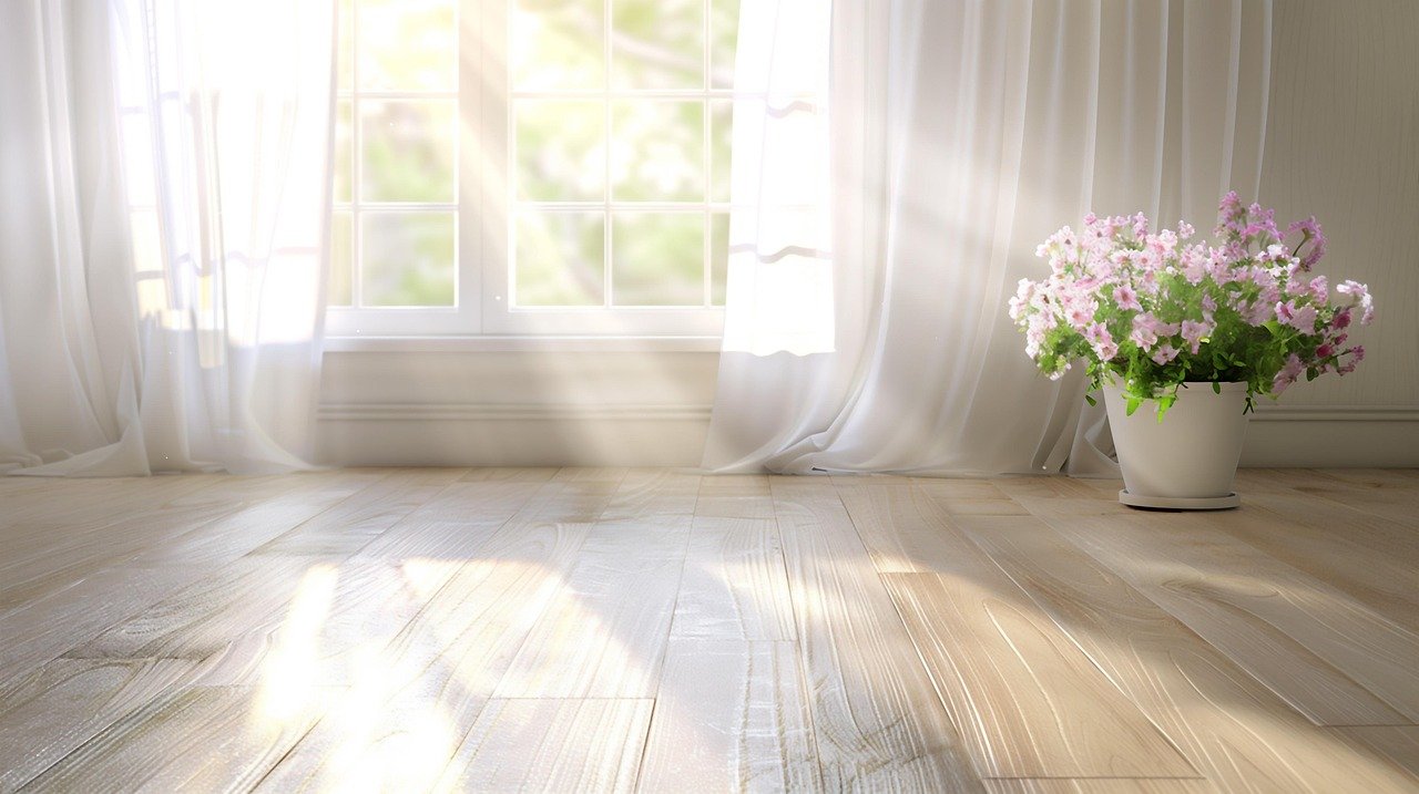 Sunlight streaming through a window with sheer curtains onto a light wooden floor, with a potted pink and purple flower plant on the right side.