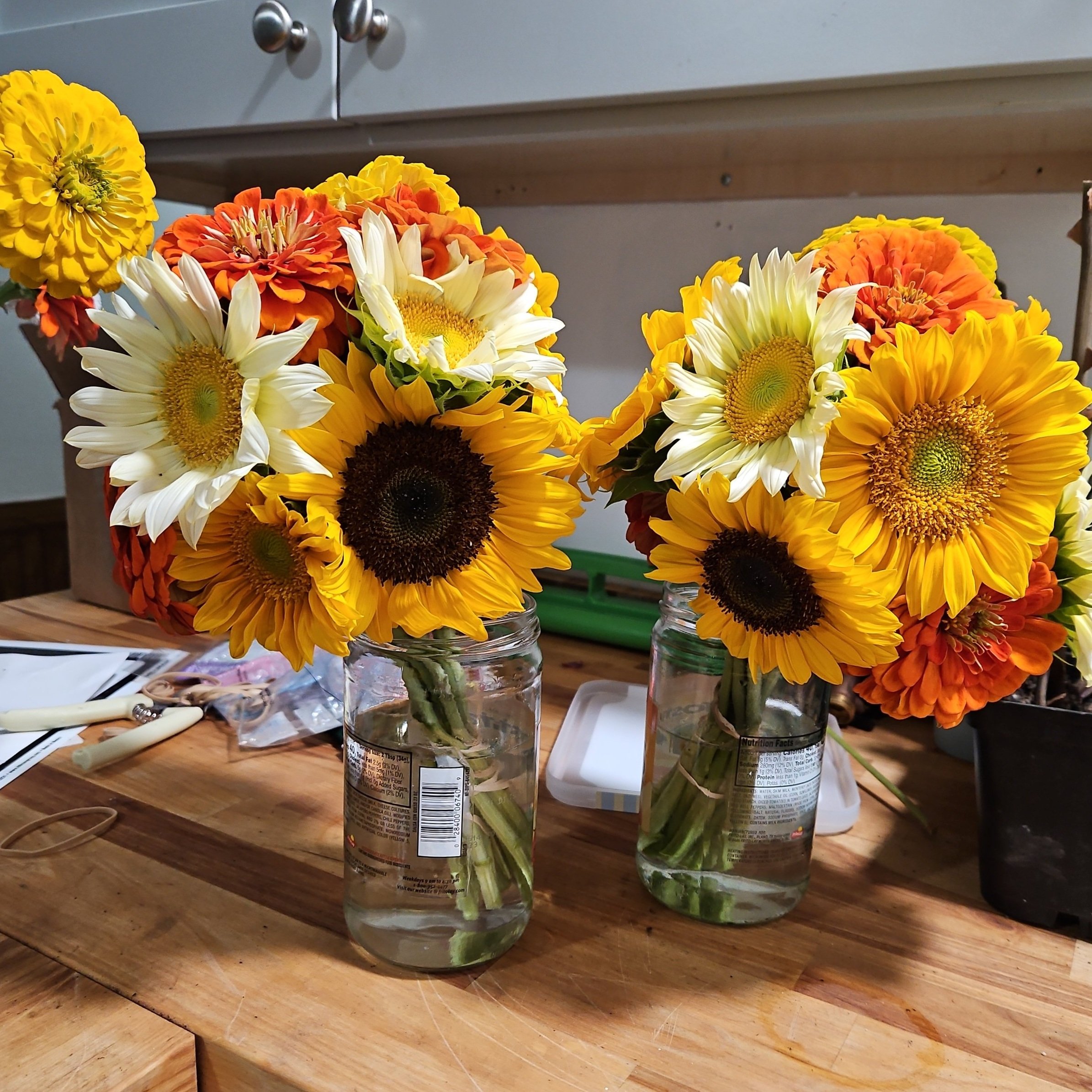 Two glass jars filled with water and arranged with colorful sunflower and zinnia flowers on a wooden table.