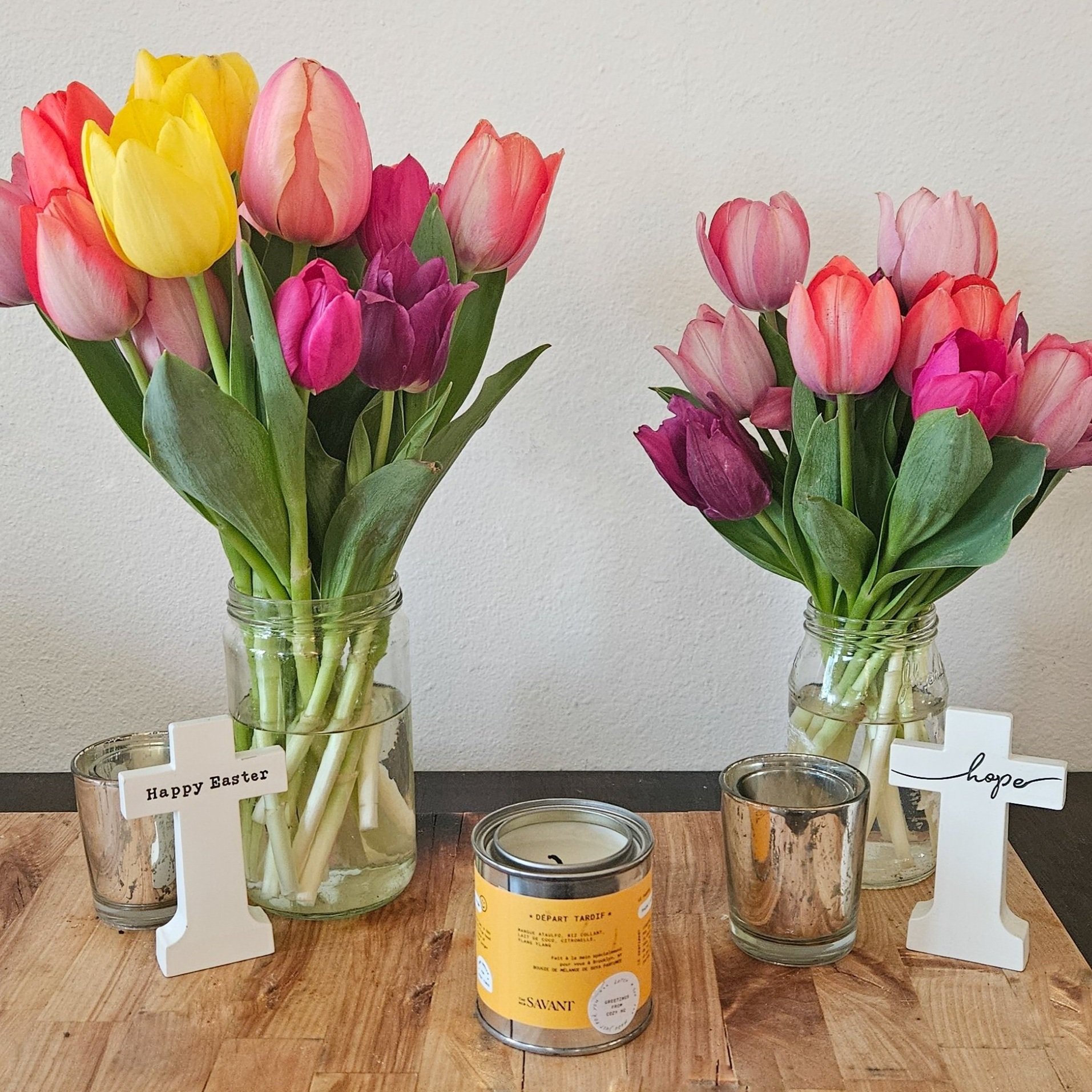 Two vases of tulips, one with yellow, pink, purple, and red flowers, and the other with pink and purple flowers, on a wooden surface. There are small signs in front of each vase saying 'Happy Easter' and 'hope'. A candle and two small glass holders a