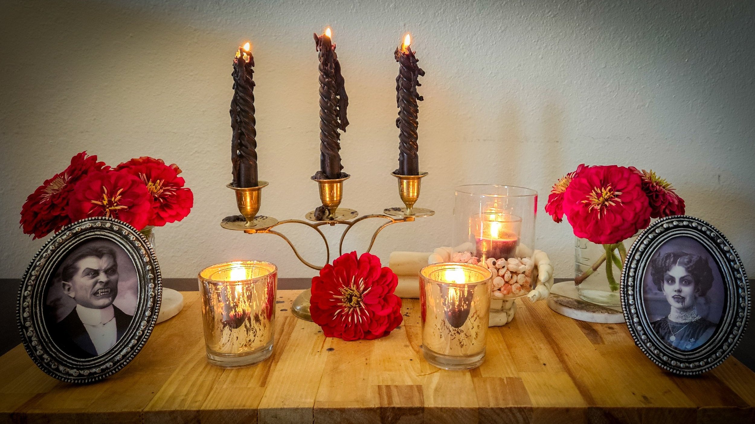 A home altar with red flowers, lit candles, framed vintage photos of women, and various decorative items.