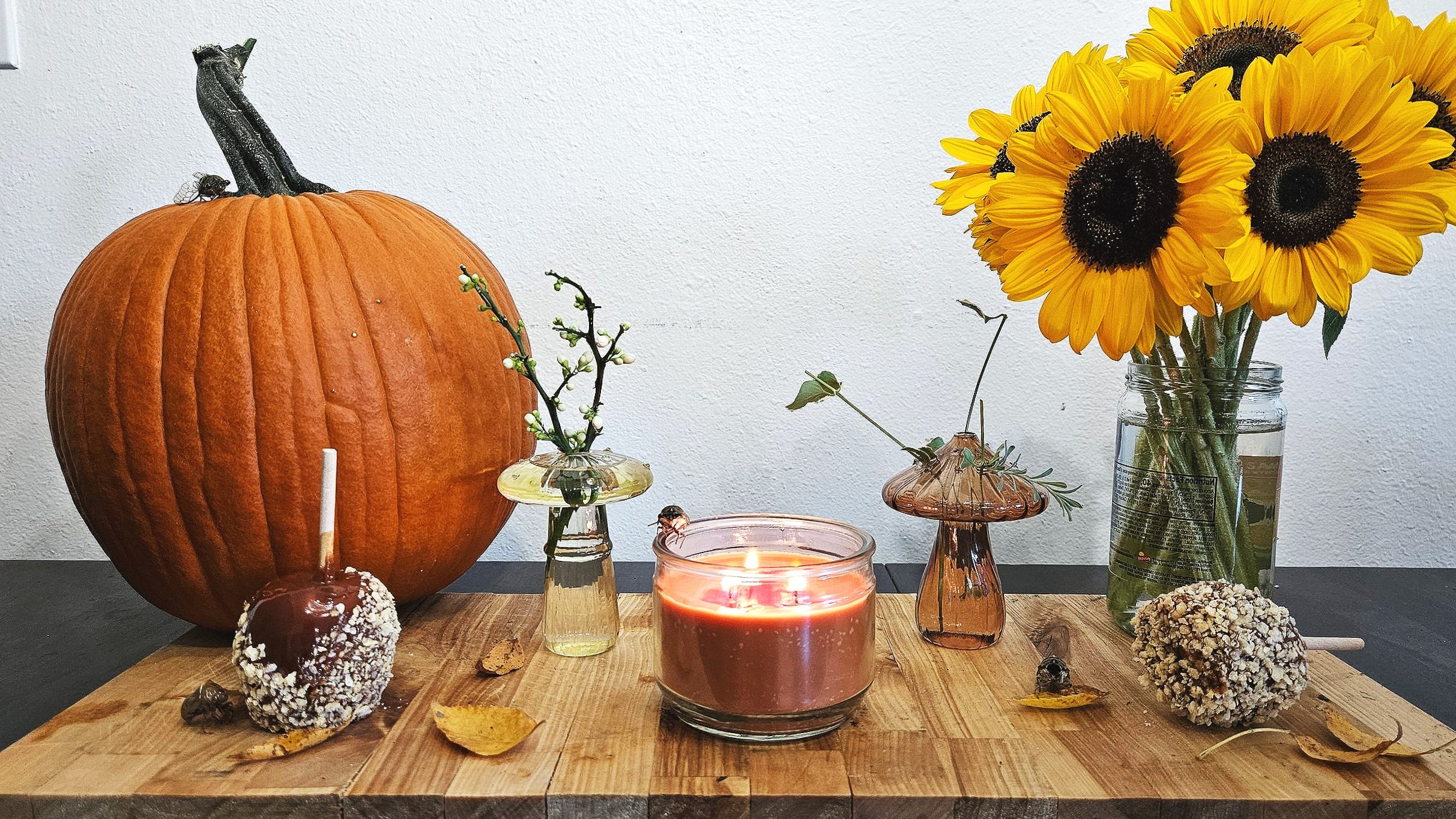 A still life arrangement on a wooden surface featuring a large pumpkin, a bouquet of sunflowers in a glass jar, a small glass vase with branches, a lit candle, and two caramel apples with white sprinkles and sticks.