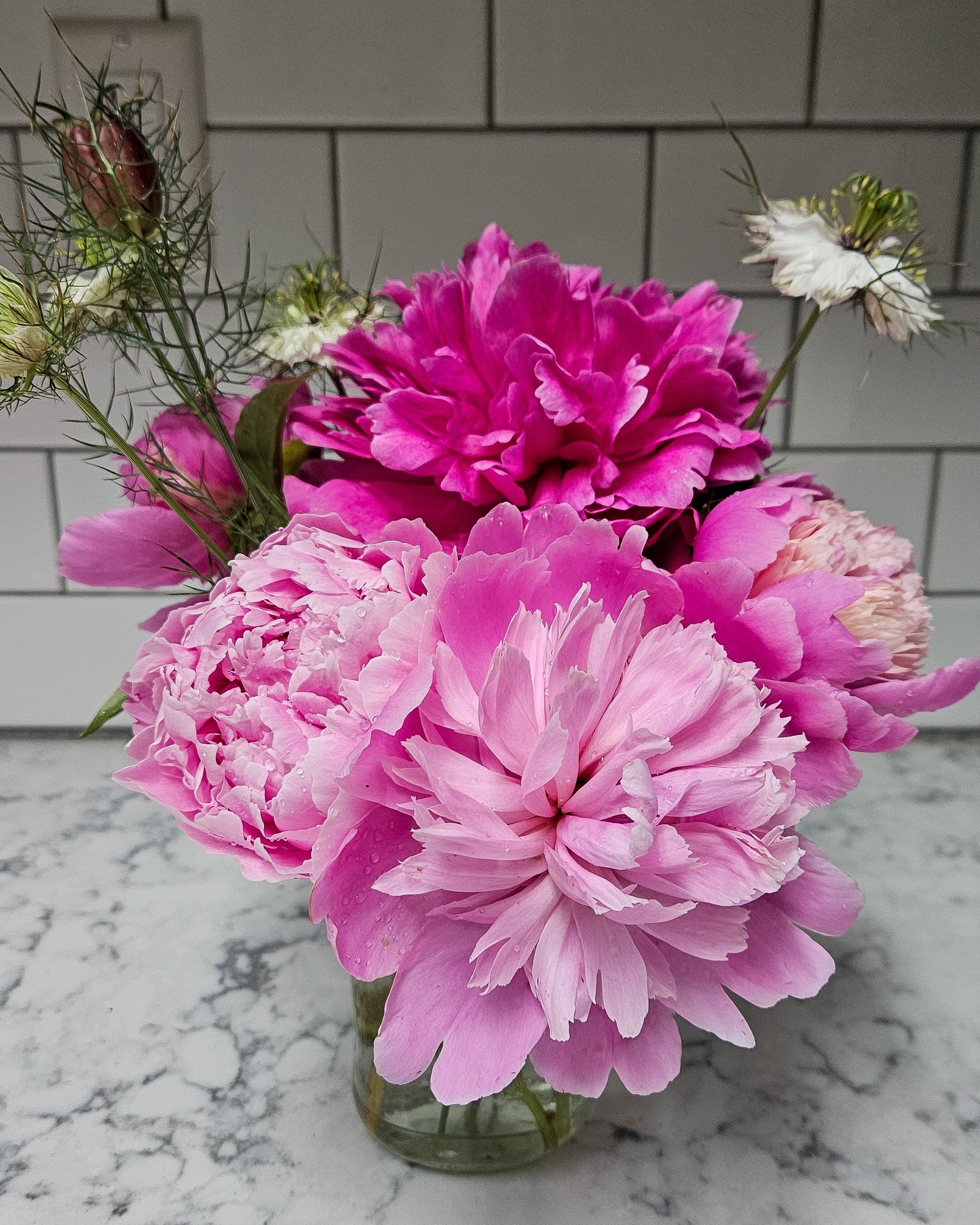 Pink peonies and white flowers in a small glass jar on a white marble surface against a tiled wall