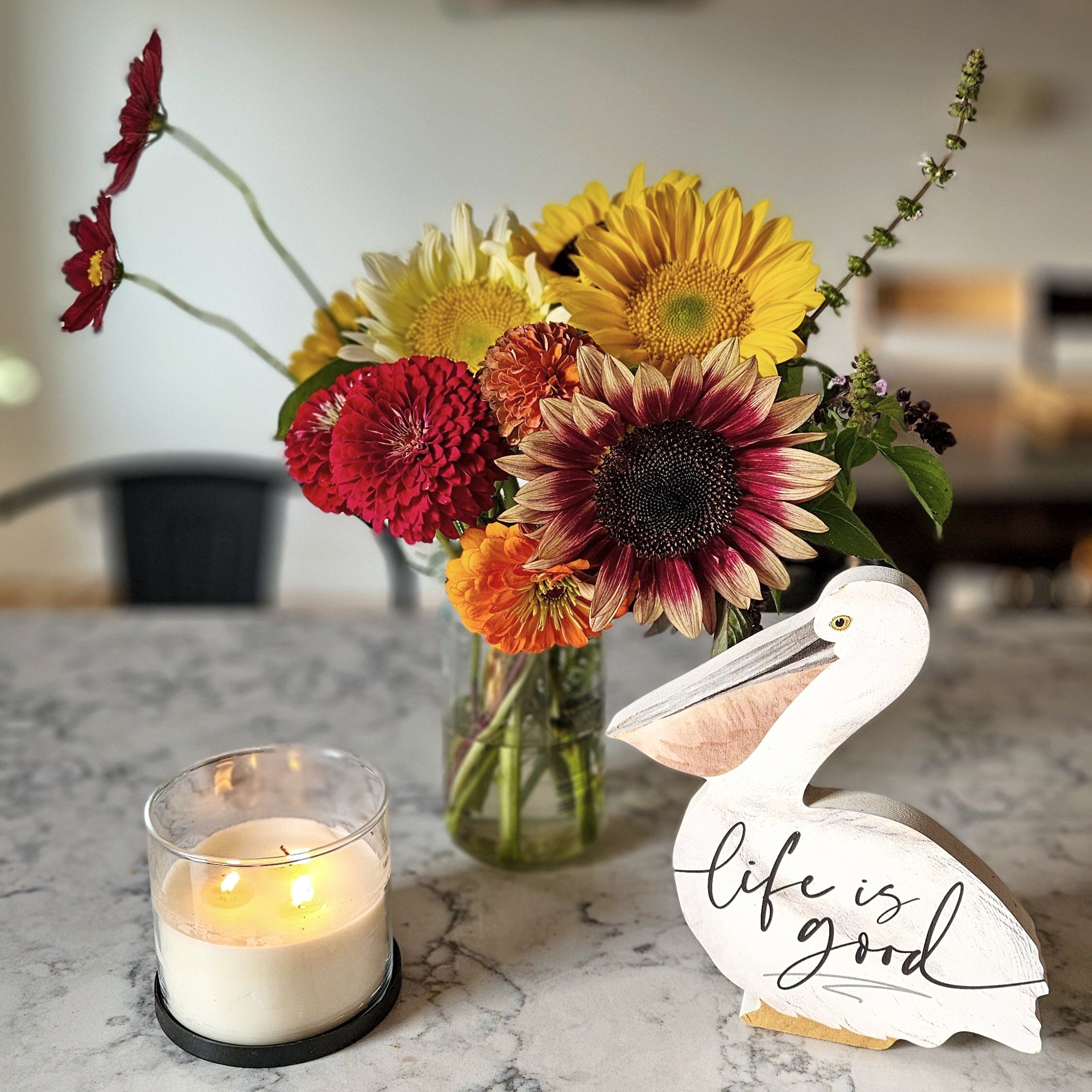 A bouquet of colorful fall flowers in a clear glass vase, a white candle in a glass holder with lit flames, and a white wooden bird decoration with the words "life is good" written on it, all arranged on a marble countertop.