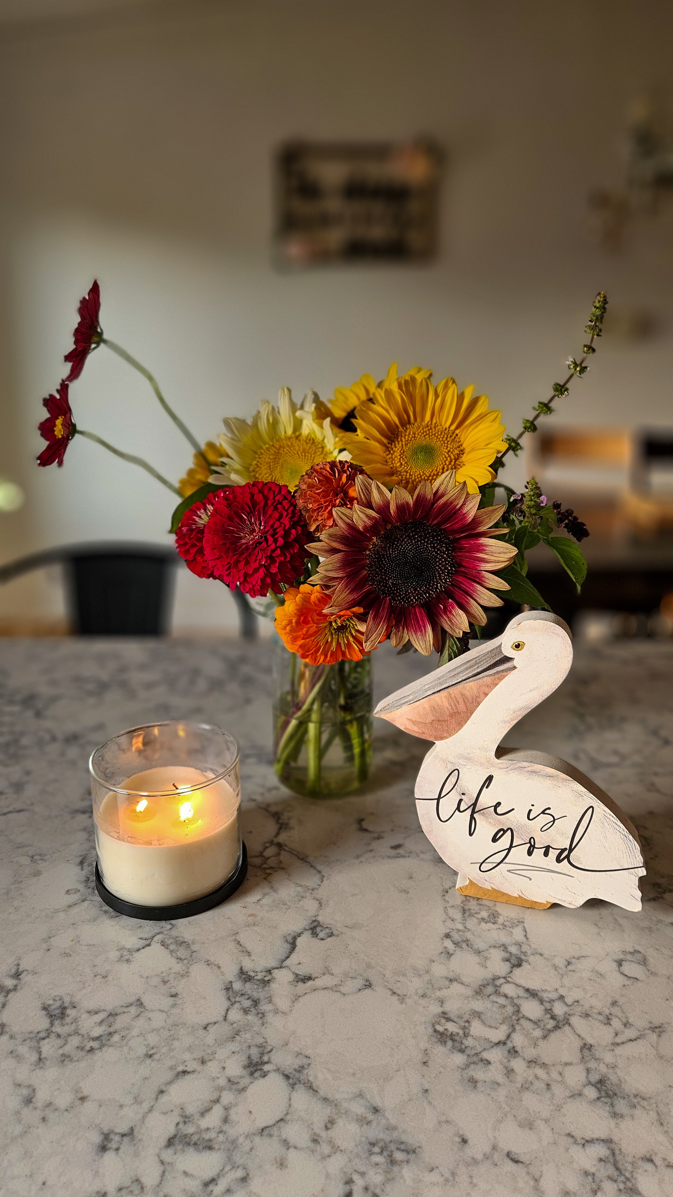 A marble kitchen countertop with a lit candle in a glass holder, a vase of colorful flowers, and a wooden pelican decoration with the words "life is good" written on it.