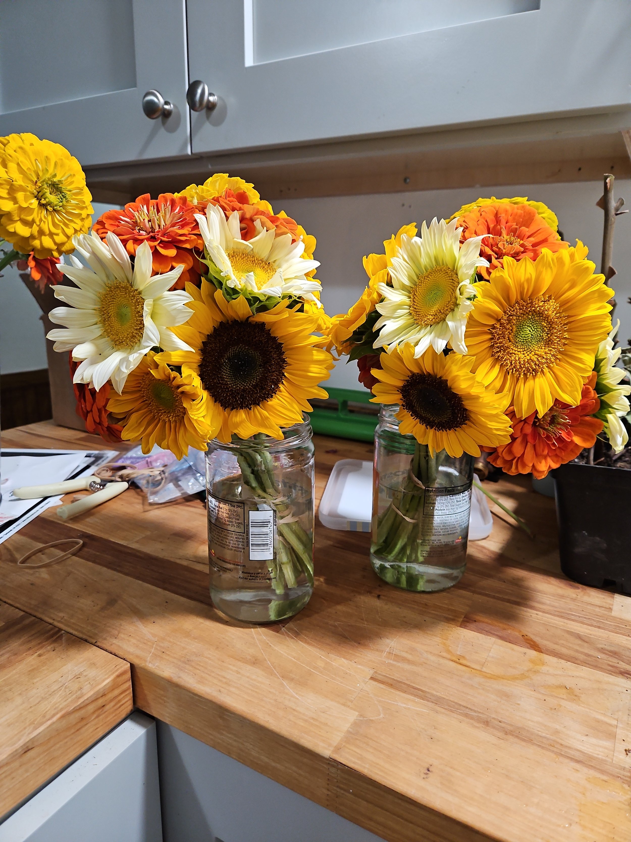 Two mason jars with sunflowers, zinnias, and marigolds on a wooden table in a kitchen.