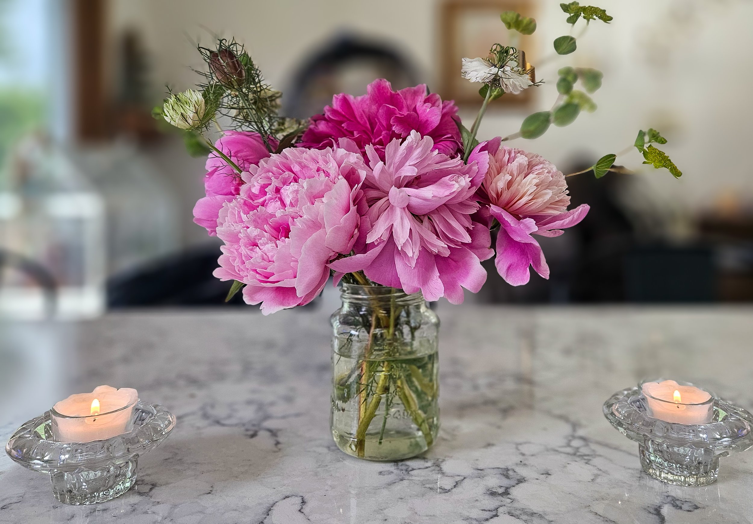 Pink peony flowers in a glass jar on a marble table with two lit candles in glass holders.