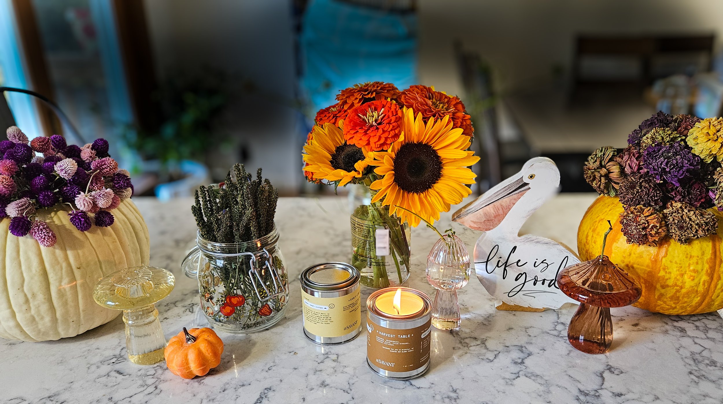 Decorative autumn-themed table centerpiece with pumpkins, sunflowers, dried flowers, candles, and glass ornaments on a marble surface.