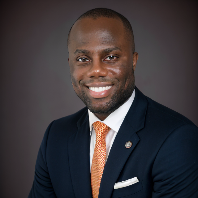 Professional portrait of a smiling African American man in a navy suit, white shirt, and orange tie, against a dark background.