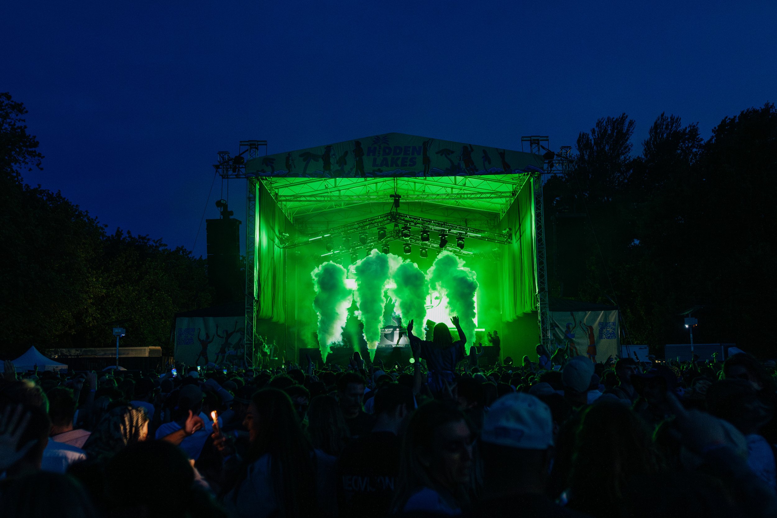Outdoor festival concert at night with green lights and smoke on stage, crowd in foreground