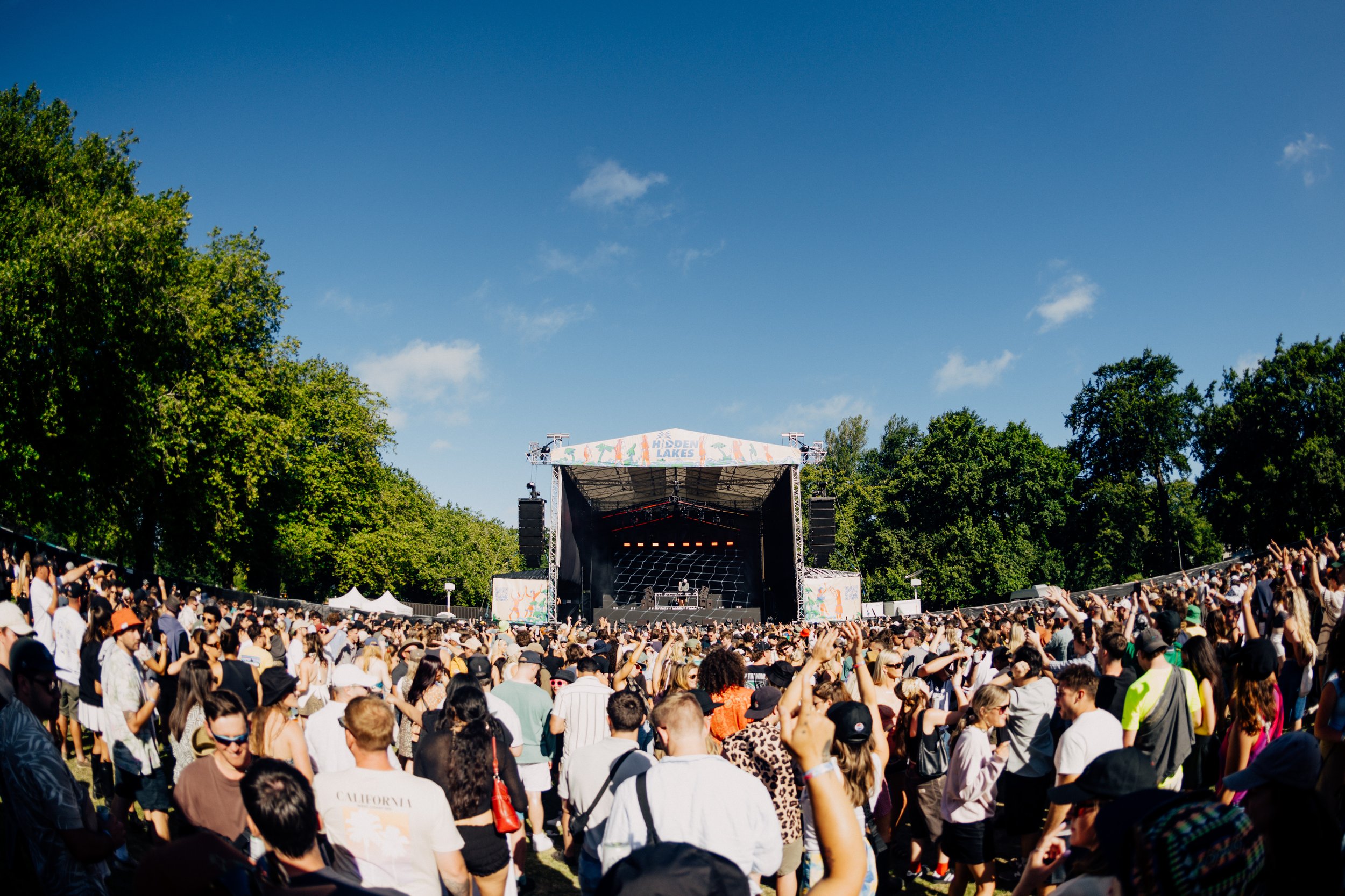 Outdoor music festival with crowd in front of a stage, labeled 'Hidden Lakes,' surrounded by trees under a clear blue sky.