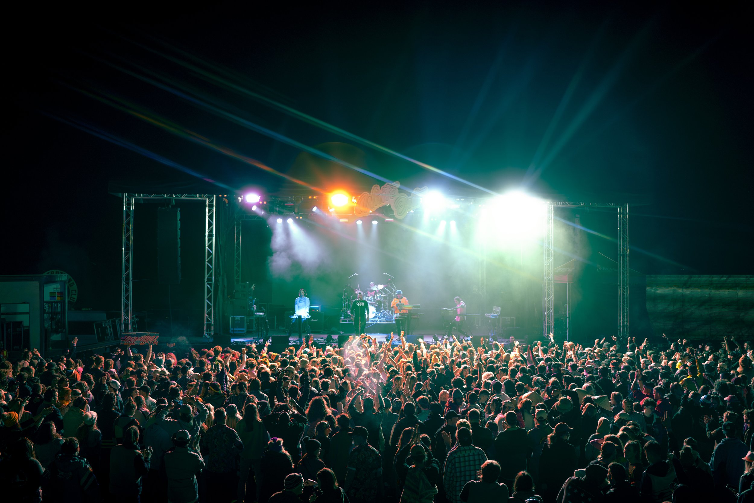 Crowd at an outdoor concert watching a band perform on a brightly lit stage at night.