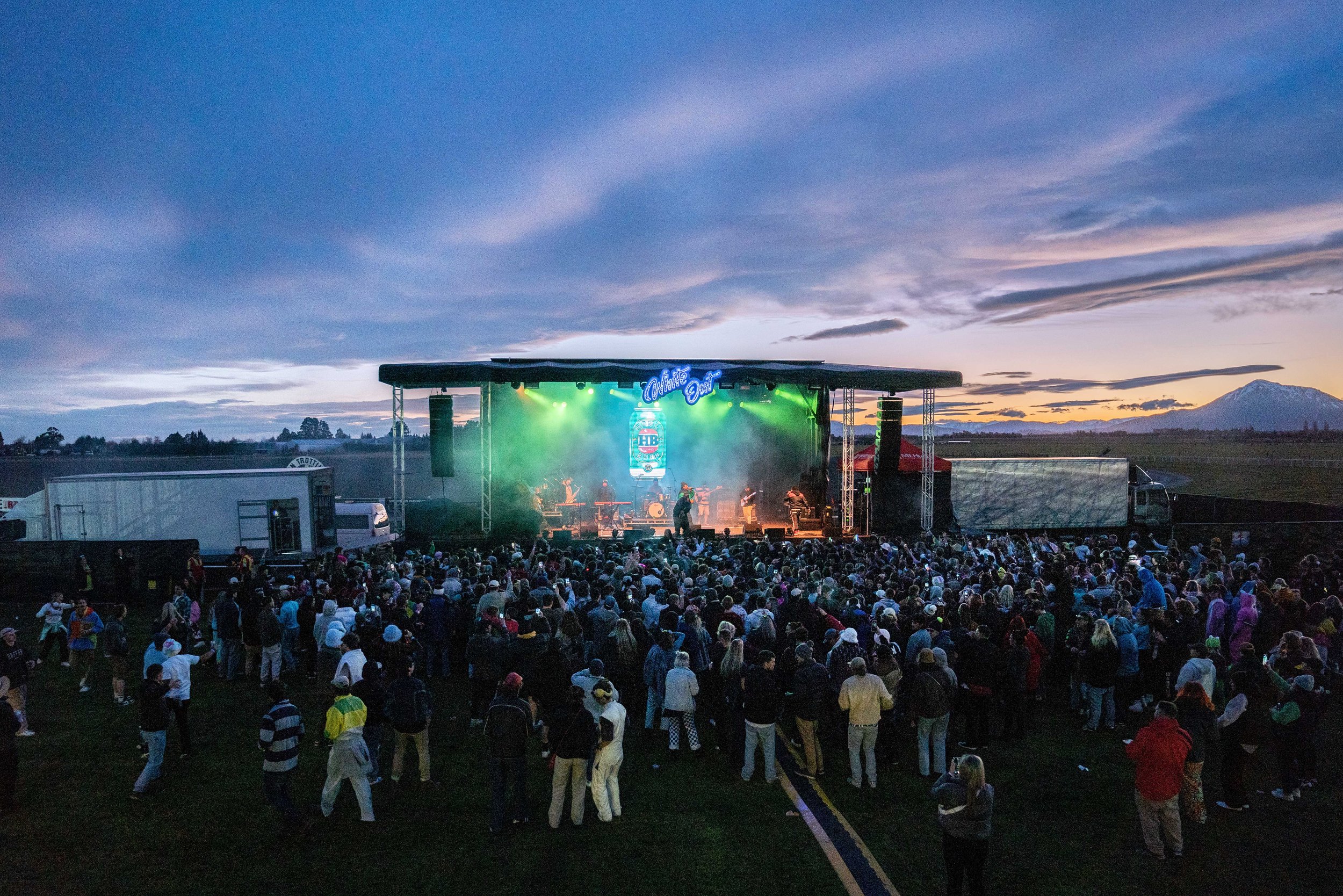 Outdoor music festival with stage, band performing, and large crowd of people, evening sky in background.
