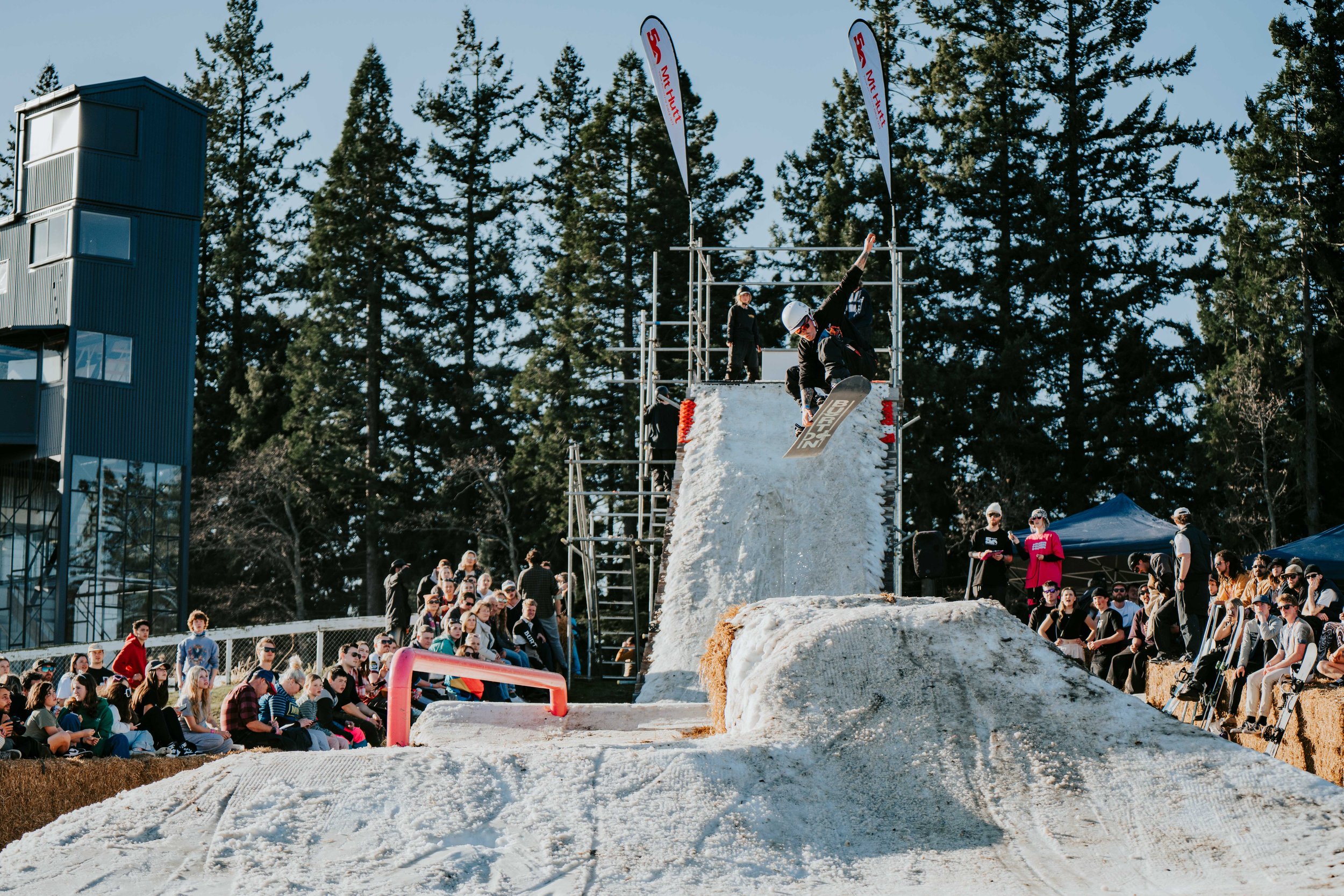 Snowboarder performing trick at outdoor event with audience watching, pine trees in background.