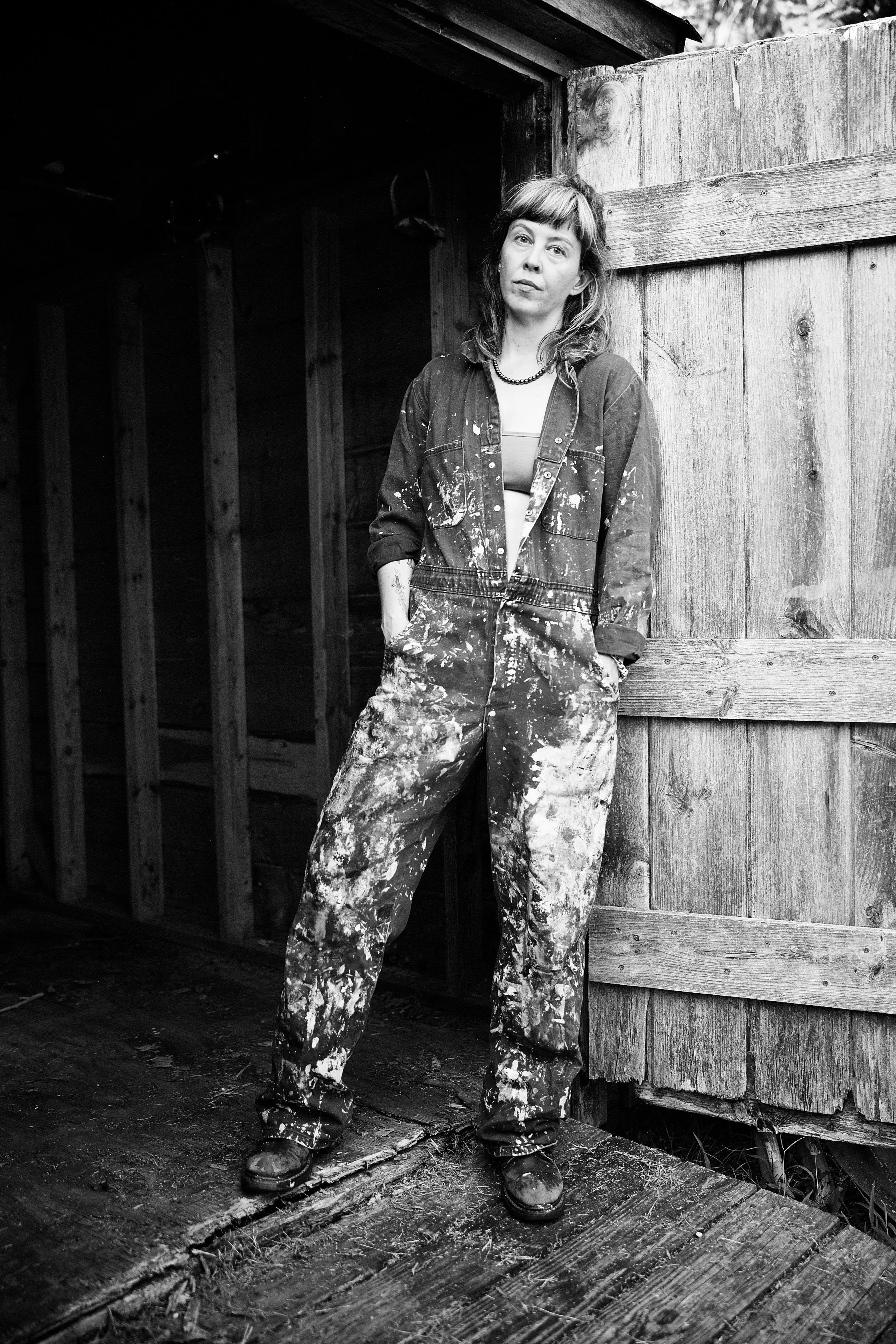 Artist Savannah C Green stands against a wooden shed with her hands in her pockets, wearing paint-stained work overalls and boots in a rustic setting.