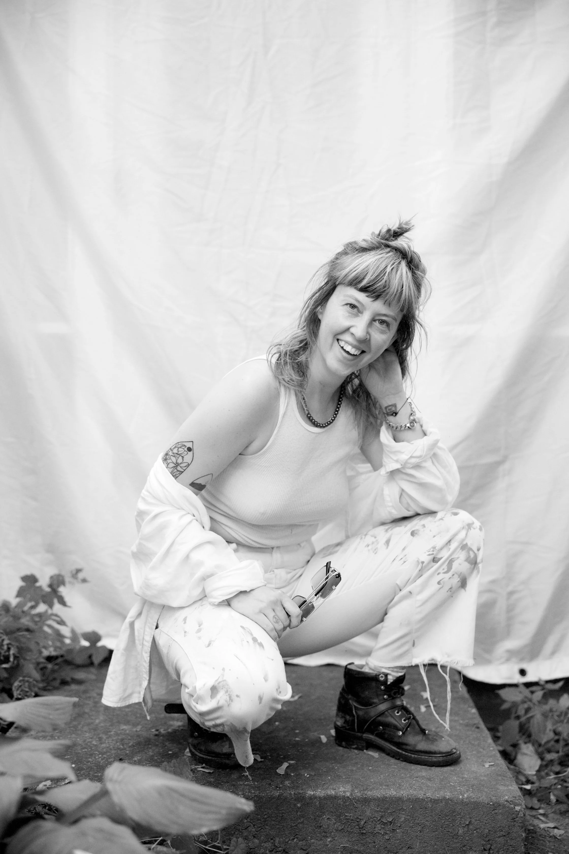 Artist Savannah C Green sitting outside on a concrete block, smiling at the camera, holding a pair of glasses. She is wearing a sleeveless top, paint-stained pants, and boots. Her hair is messy, and she has tattoos, a necklace, and bracelets.