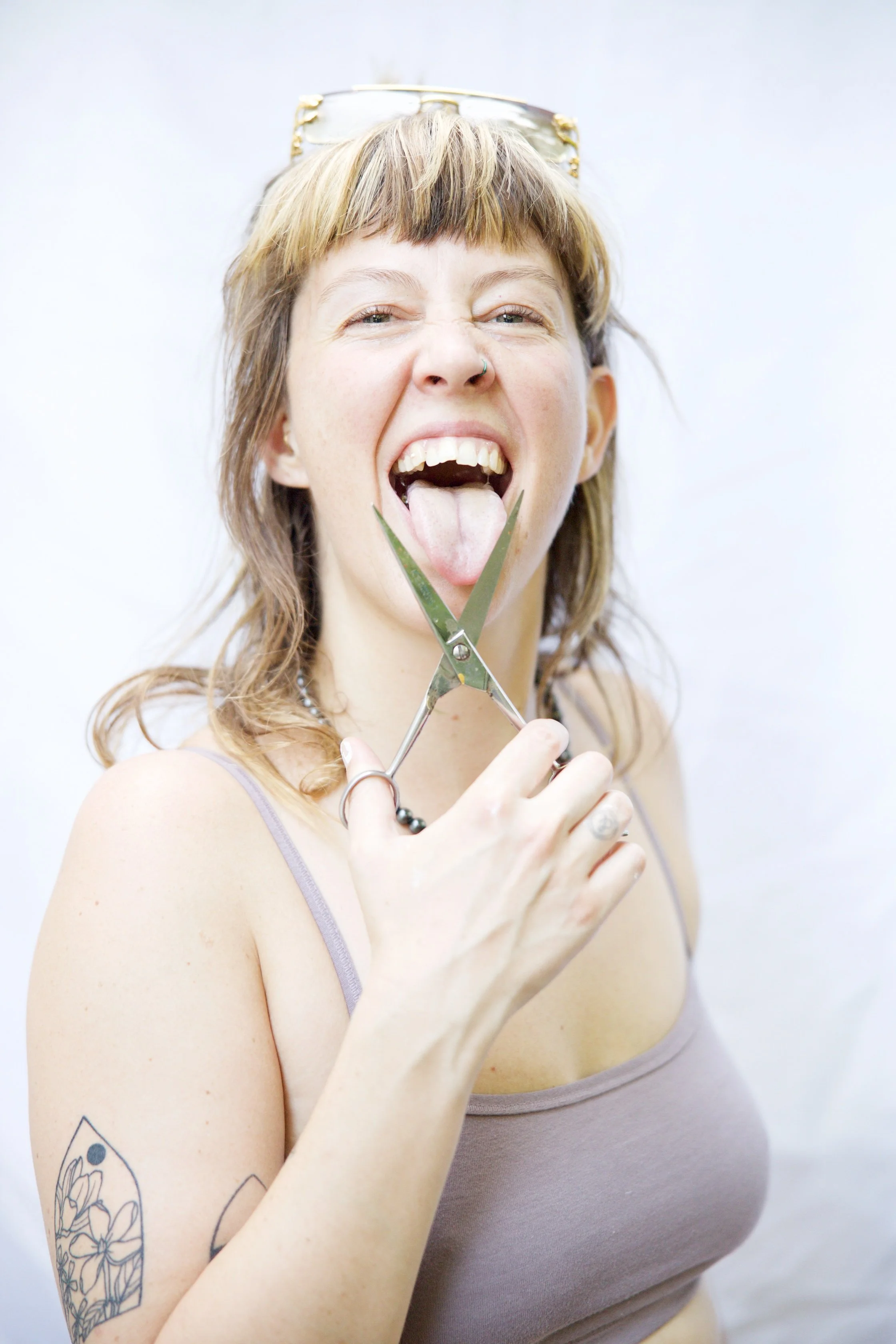 Artist Savannah C Green with tattoos on her arm holding scissors near her tongue and smiling.
