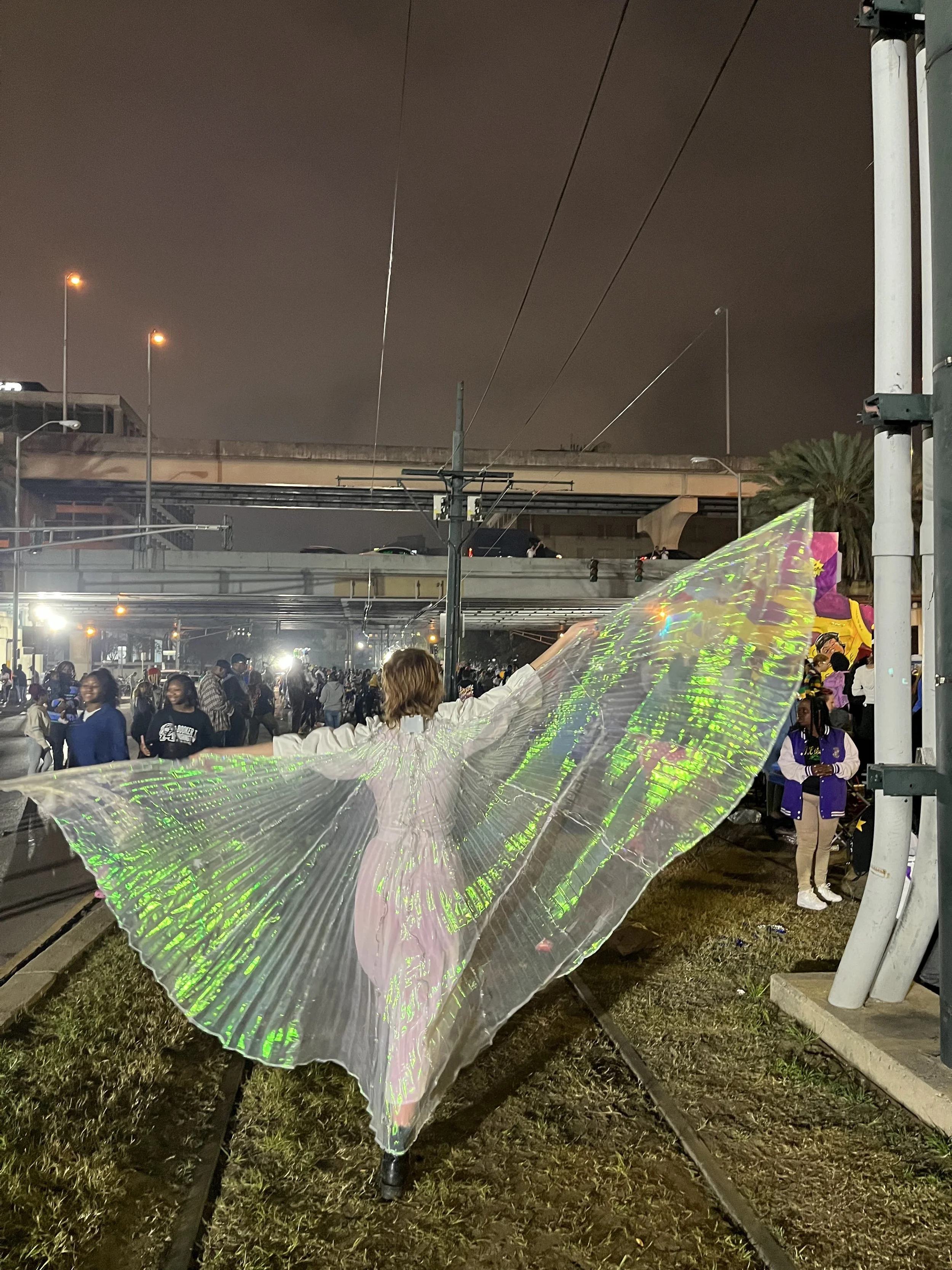 Person dressed in a white outfit with large iridescent butterfly wings standing on city street at night with people gathered in the background.