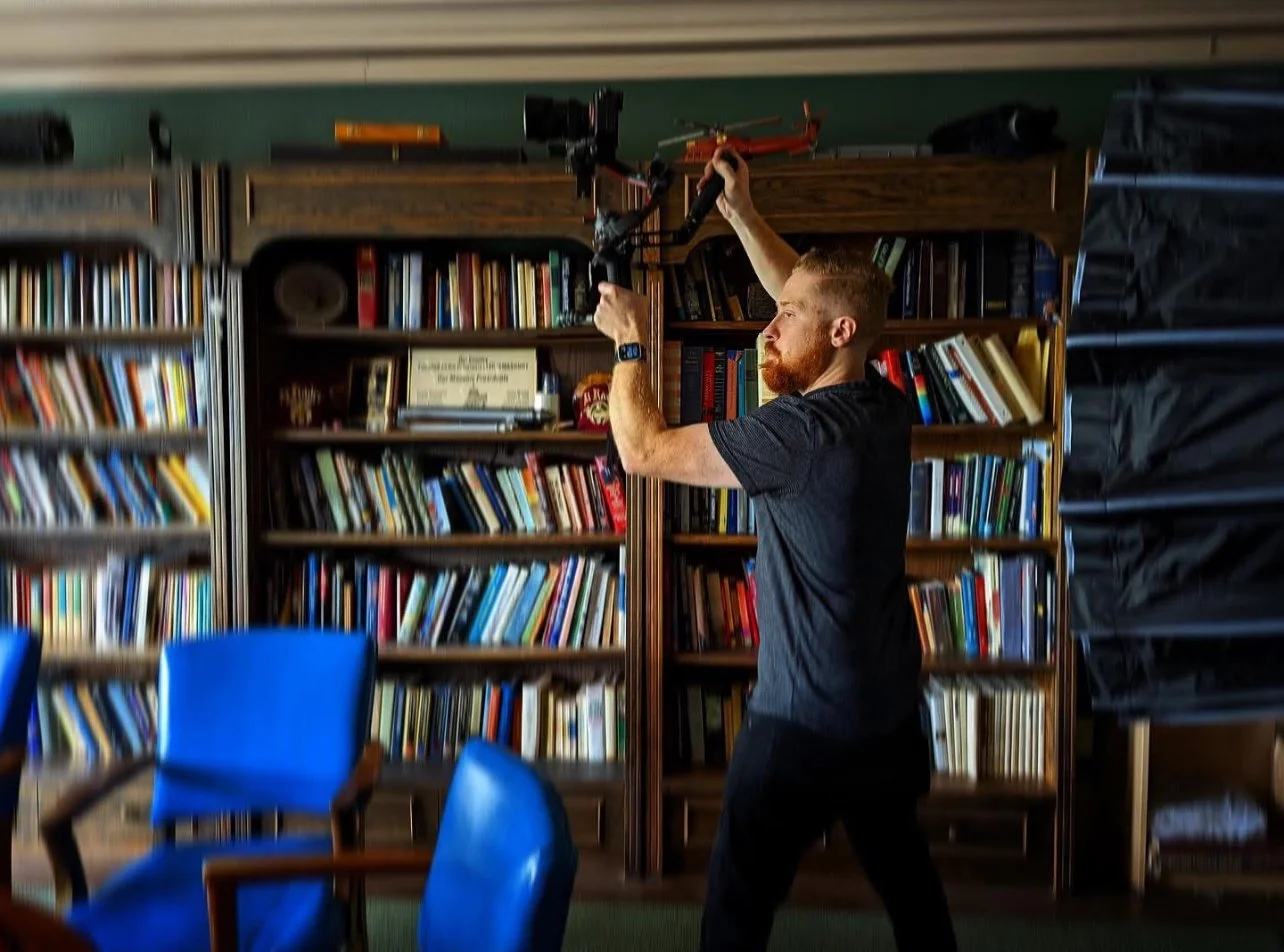 Joseph Gary adjusting professional camera gear in a creative studio setting, with bookshelves and blue chairs in the background — representing his work at Front of House Media.