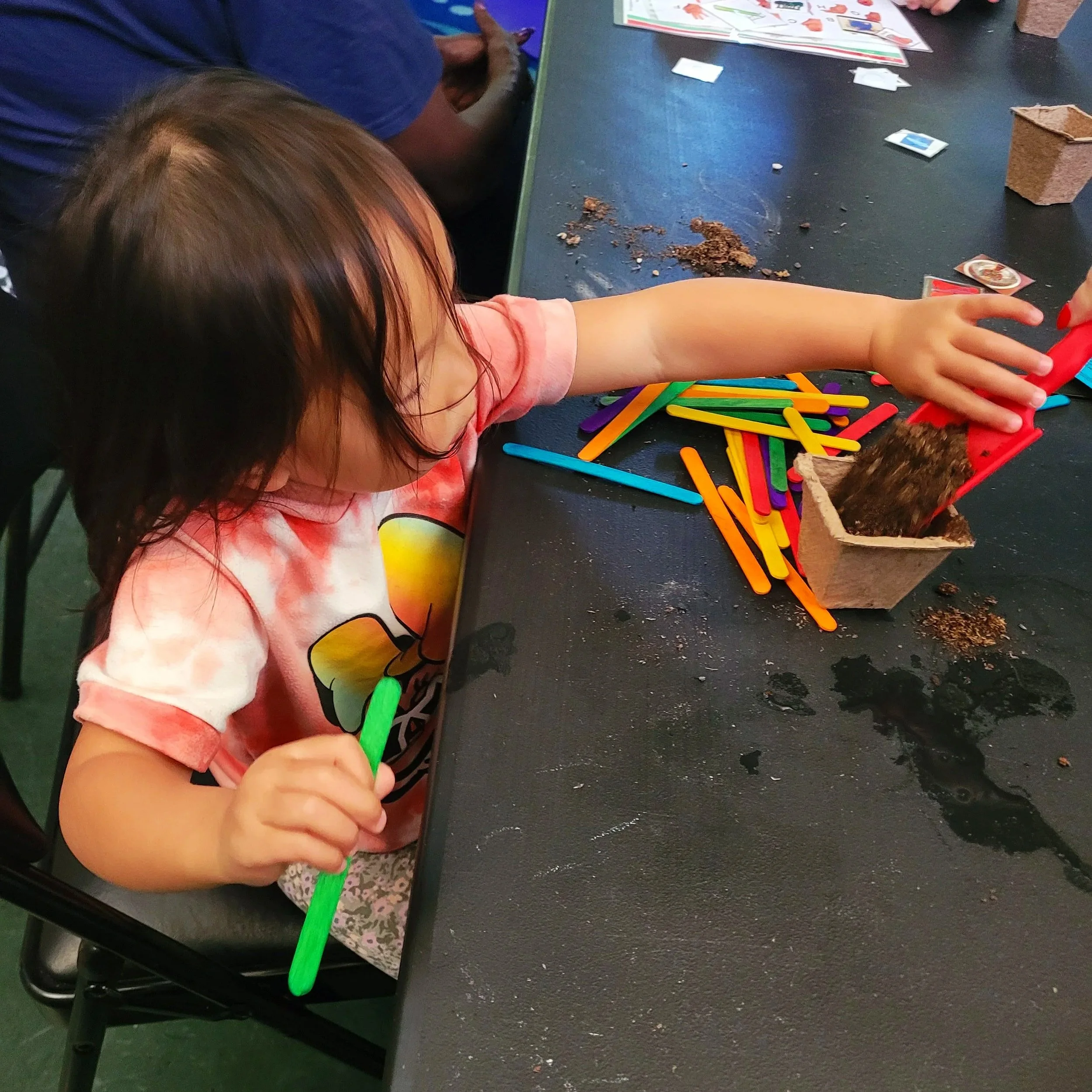 Children are playing with building blocks and colorful sticks on a black table. One child is reaching for a red stick, with other children and game pieces visible in the background.