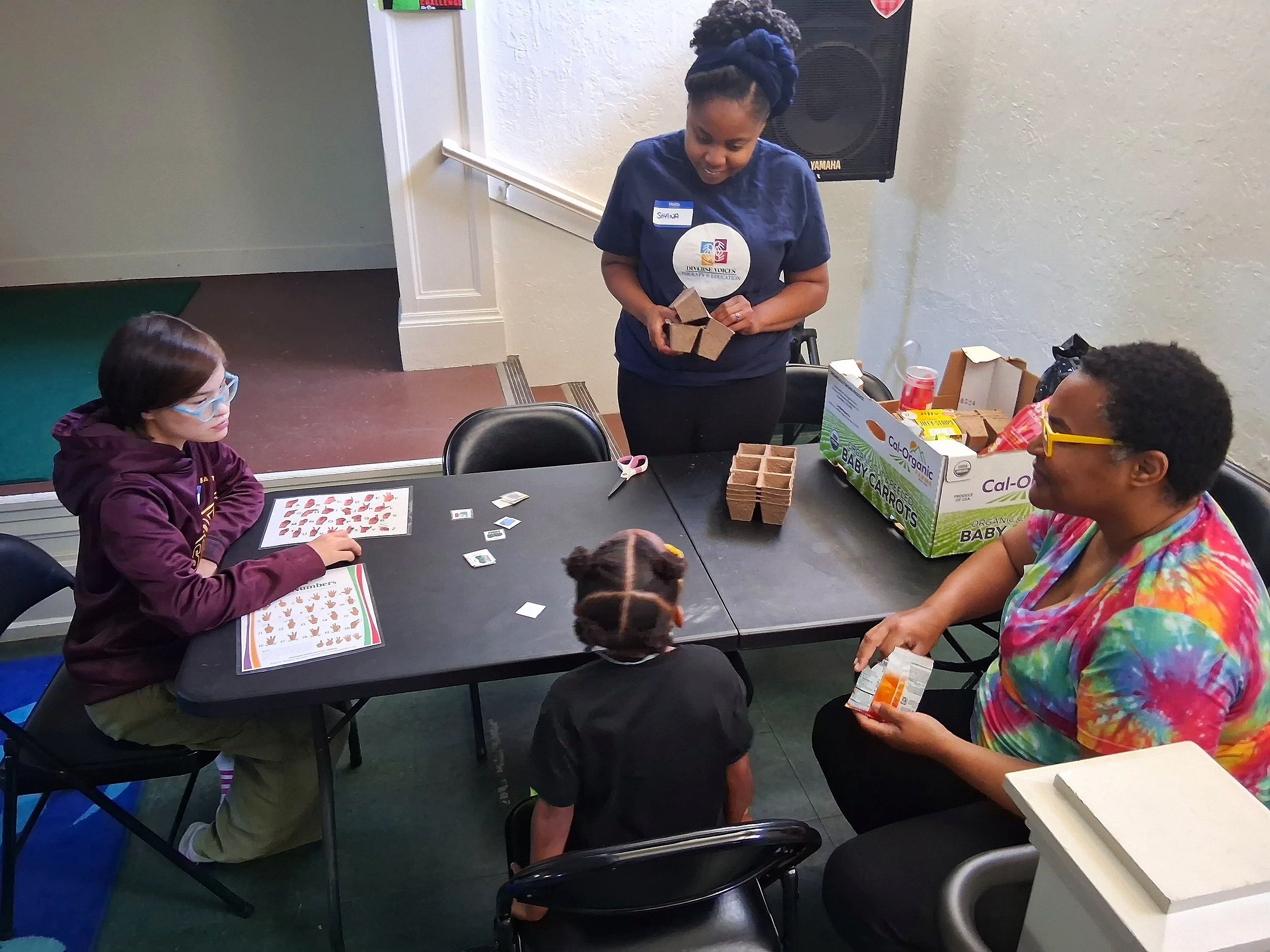 A group of children and an adult woman at a table engaged in a craft activity, with supplies like scissors, stickers, and small cardboard boxes. The woman is standing, organizing craft materials, while the children are seated, working on their projec