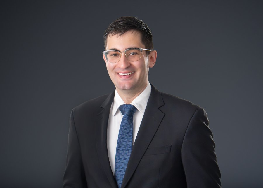 A smiling man wearing glasses, a black suit, white shirt, and blue tie against a dark background.