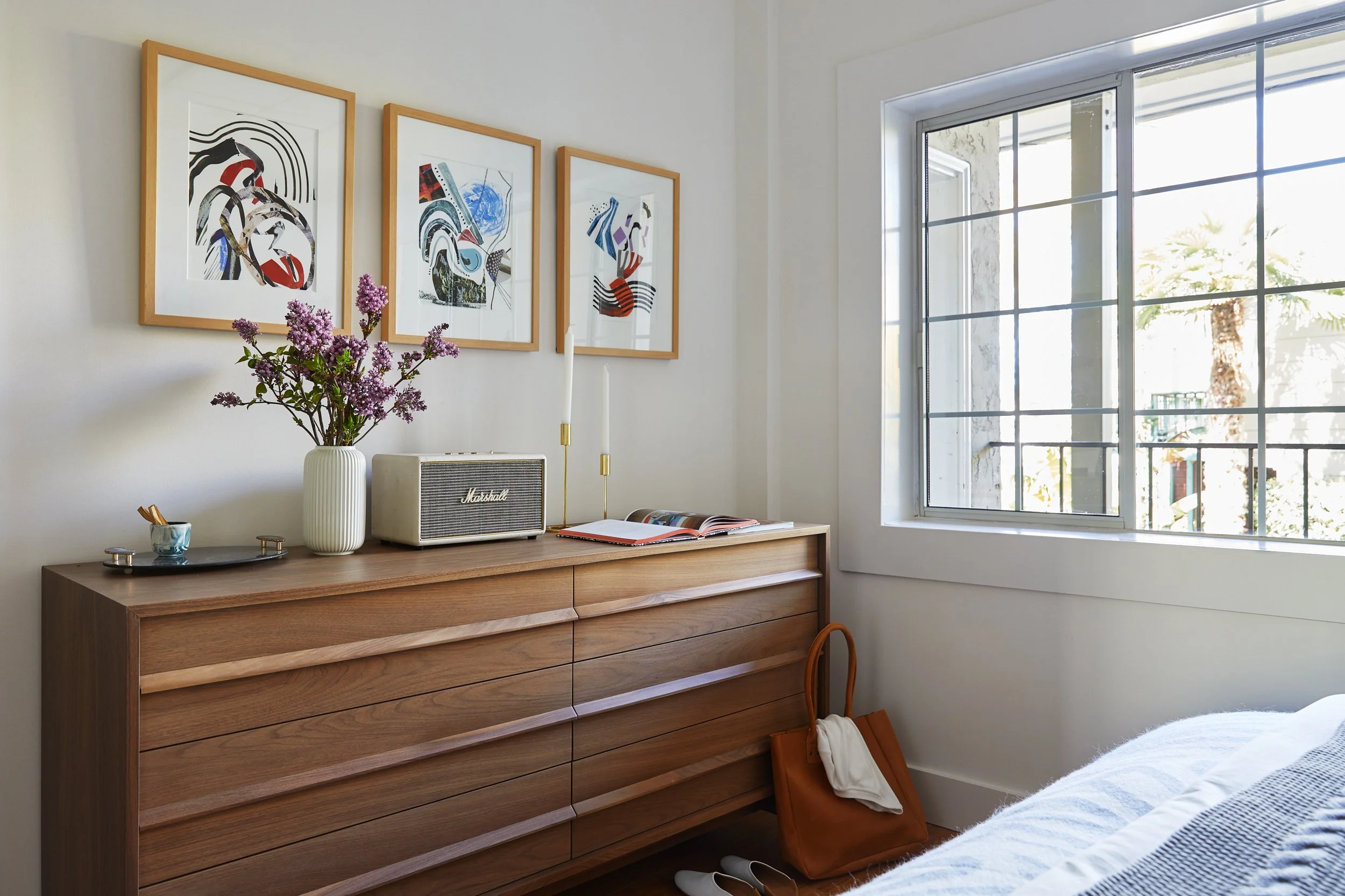 Bedroom corner with wooden dresser, abstract art on wall, vase with flowers, speaker, open book, candles, and a window with bright daylight.
