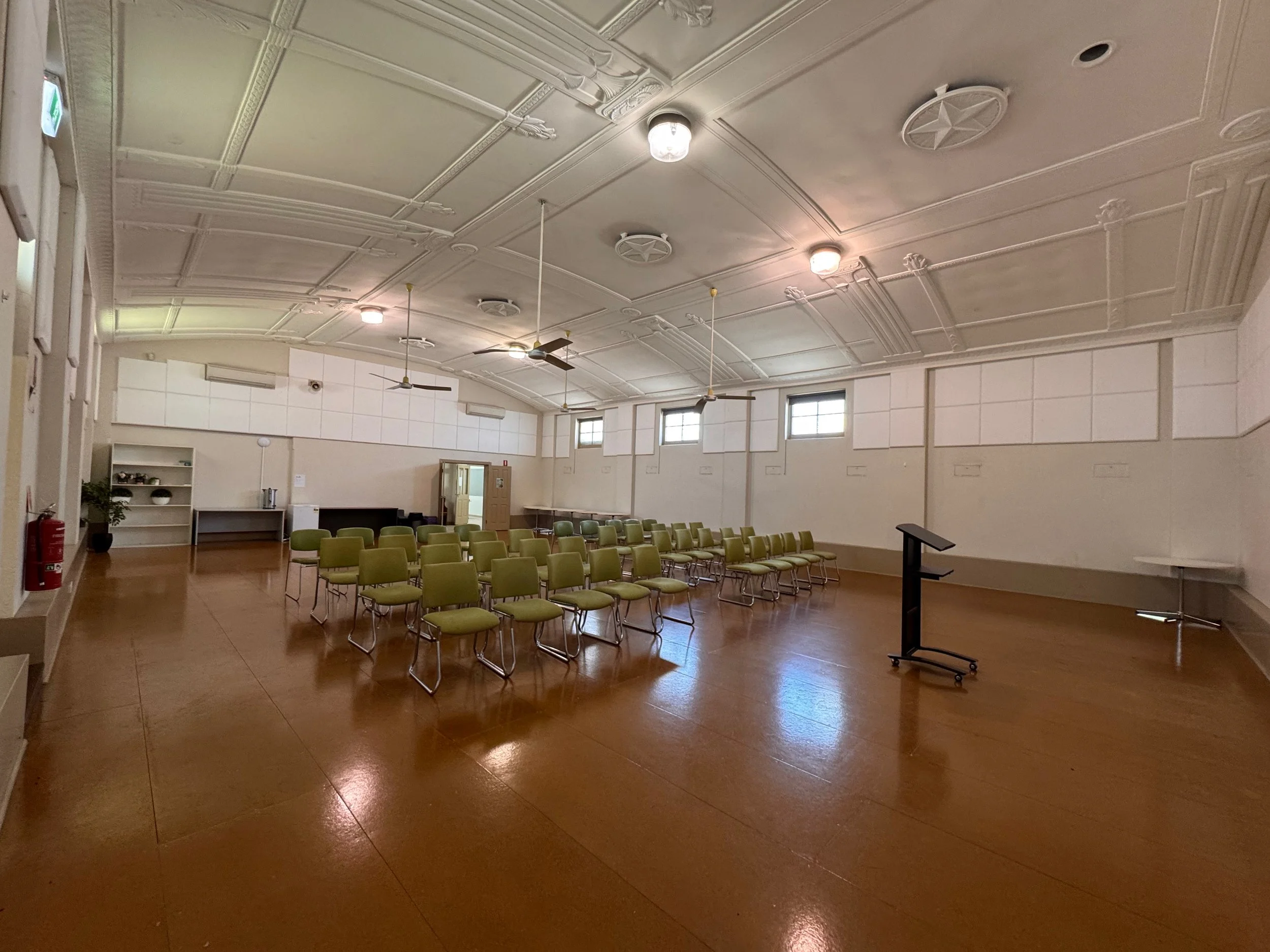 Empty auditorium with rows of green chairs, a black podium, wooden floor, and decorative ceiling, lit by overhead lights and natural light from windows.