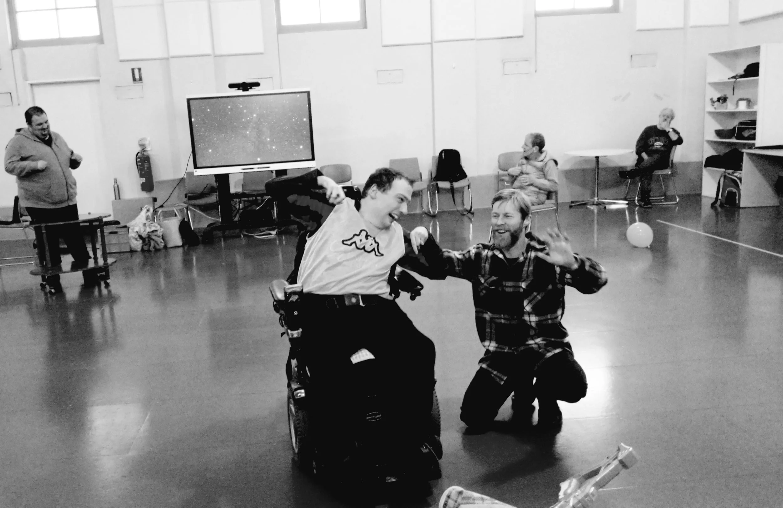 A man in a wheelchair dancing with a man kneeling in front of him, both smiling and holding hands, in a room with other seated and standing people.