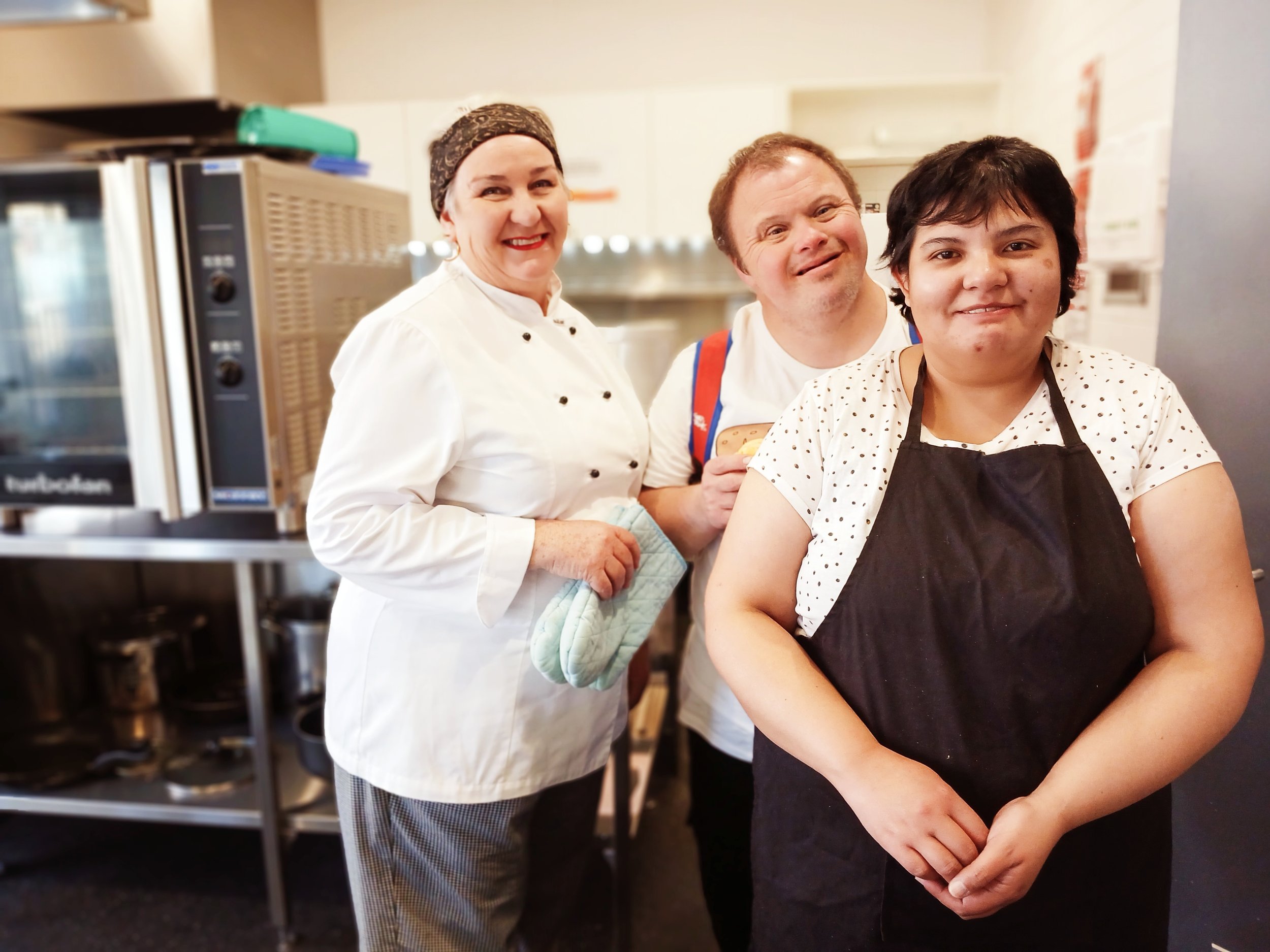 A group of three people in a kitchen, including a woman in a chef's outfit with a headscarf, a man with a backpack, and a person in a polka dot shirt and black apron, all smiling.