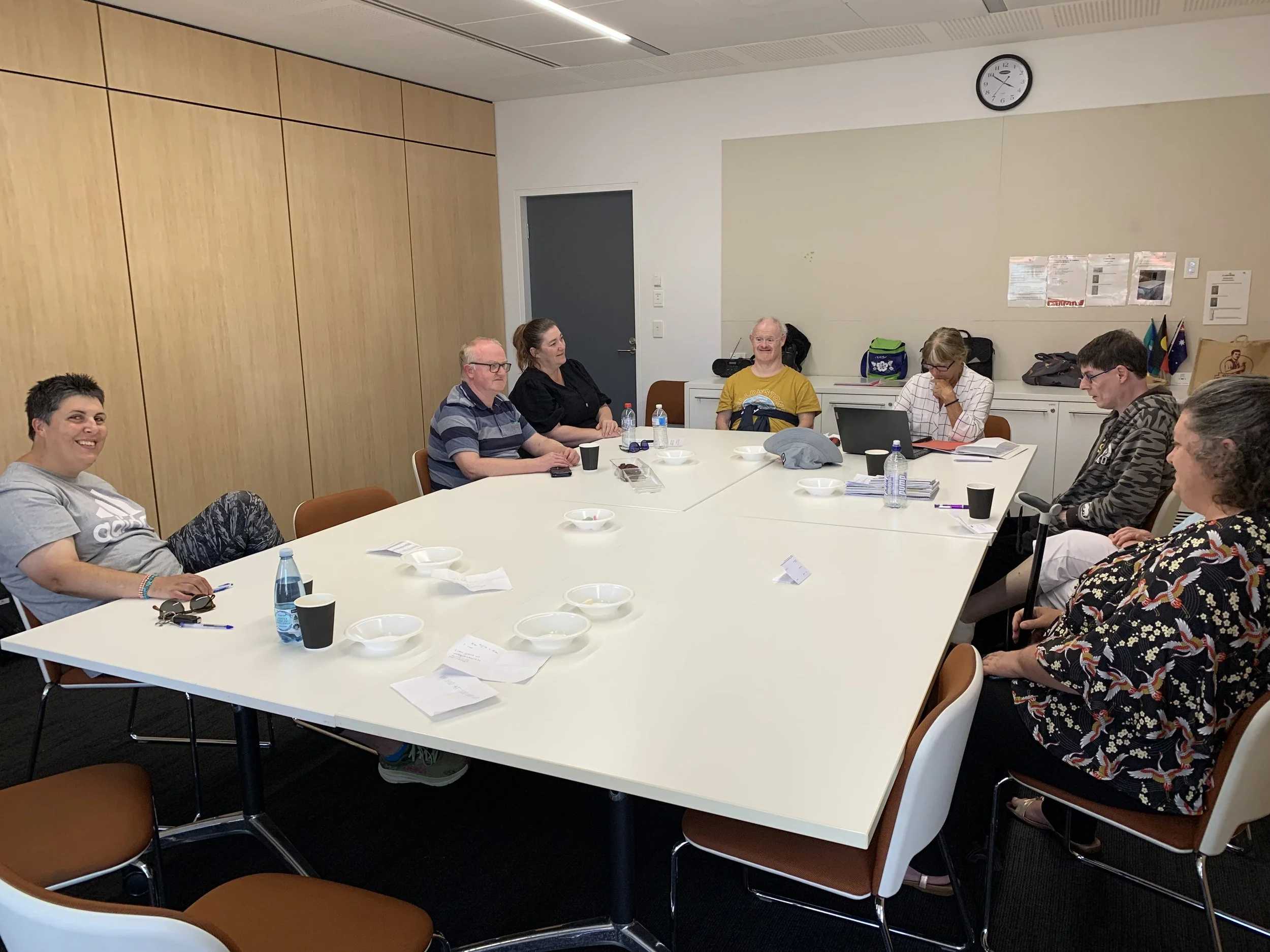 A group of eight people seated around a large white conference table in a meeting room, some smiling and engaging, with papers, water bottles, and cups on the table.
