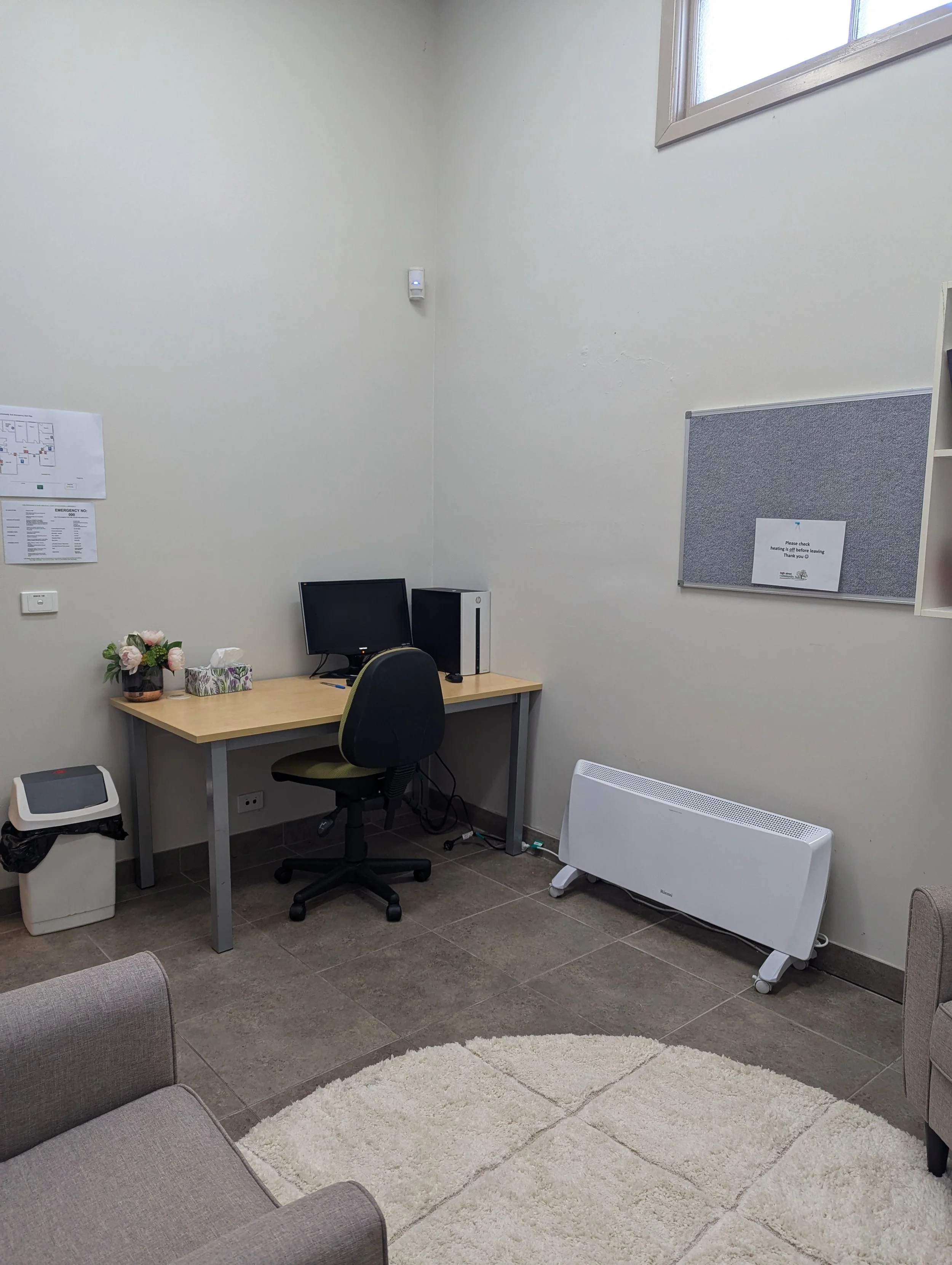A small office with a desk, computer, tissues, and flowers, a chair, a white heater, a corkboard, and a trash bin.