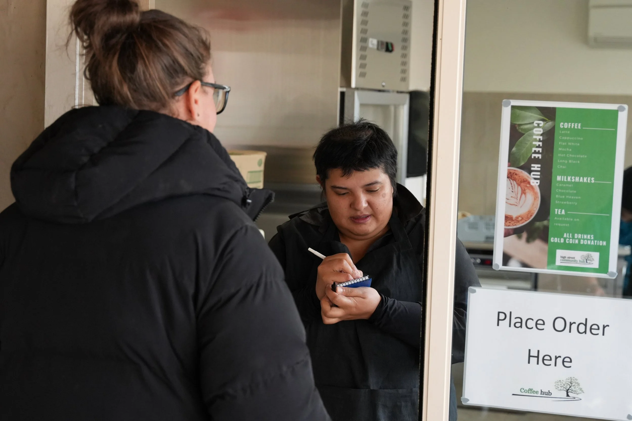 Person taking order at a cafe counter with another person standing in front, menu visible on the wall.