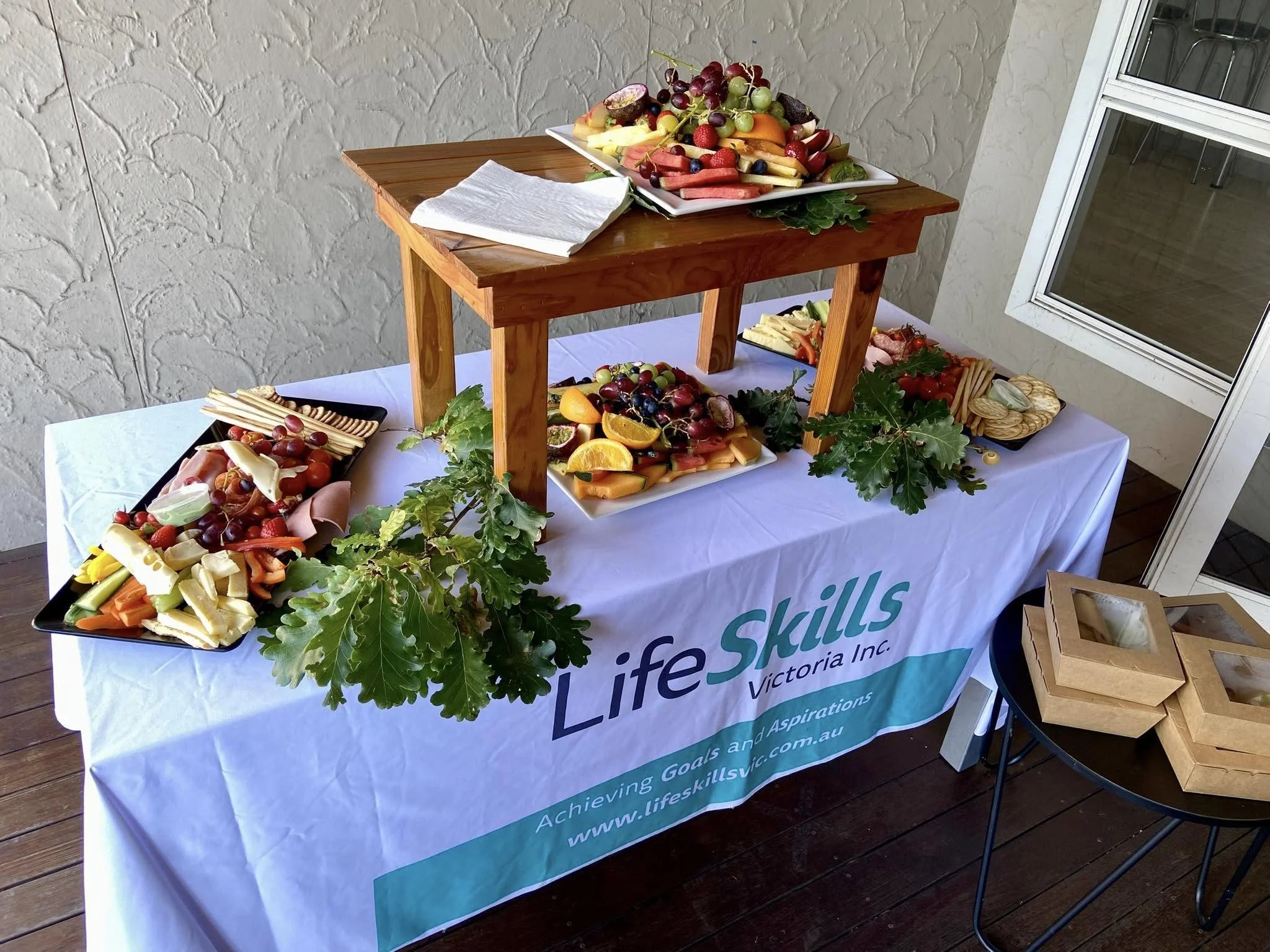 Assorted fruit and cheese platters displayed on a table with a white tablecloth. The tablecloth features the logo "LifeSkills Victoria Inc." and a website address. The setup includes a small wooden table for elevation. Boxes are nearby on a black round table.